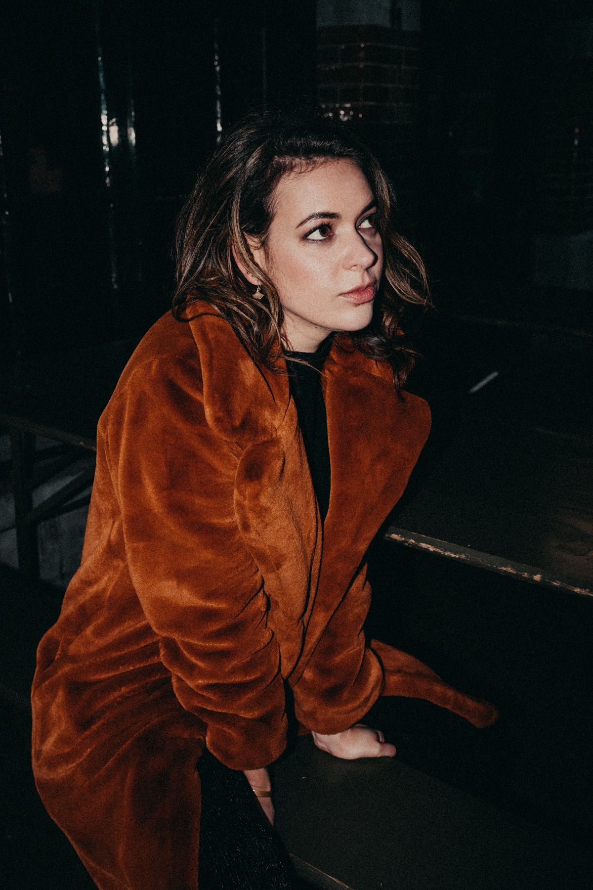 A person wearing a rust-colored velvet jacket poses in a dark moody setting with dramatic lighting.