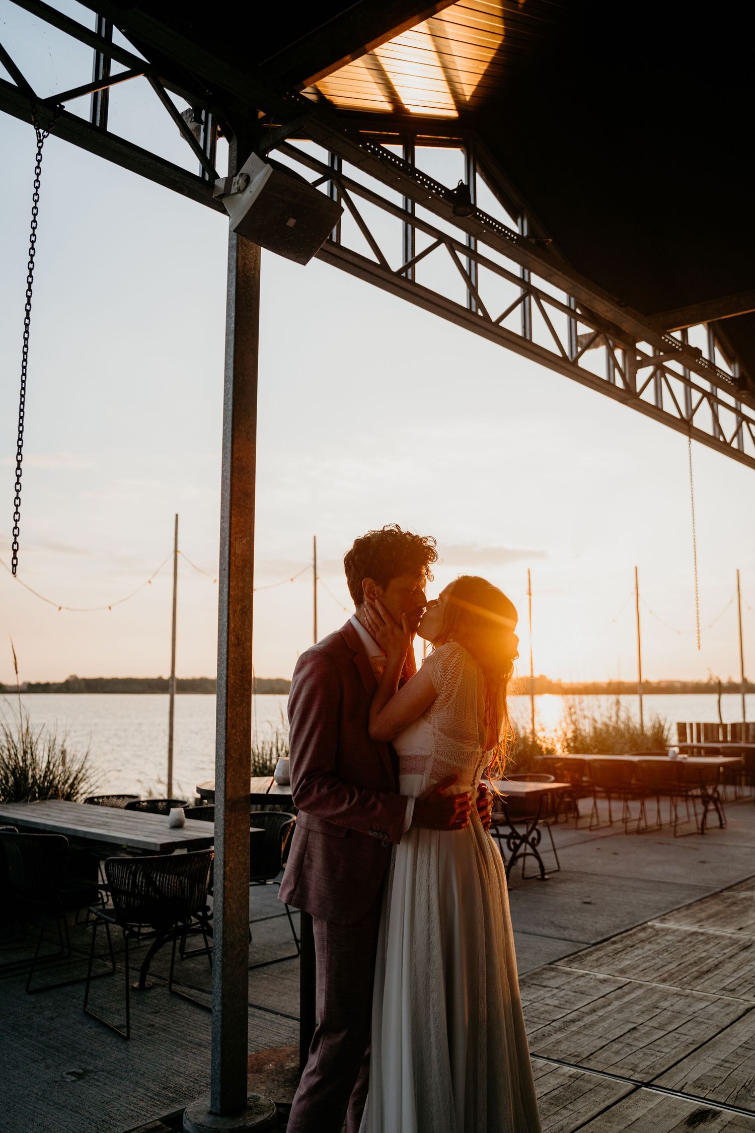 Wedding couple embracing under a covered pier at sunset with marina views.