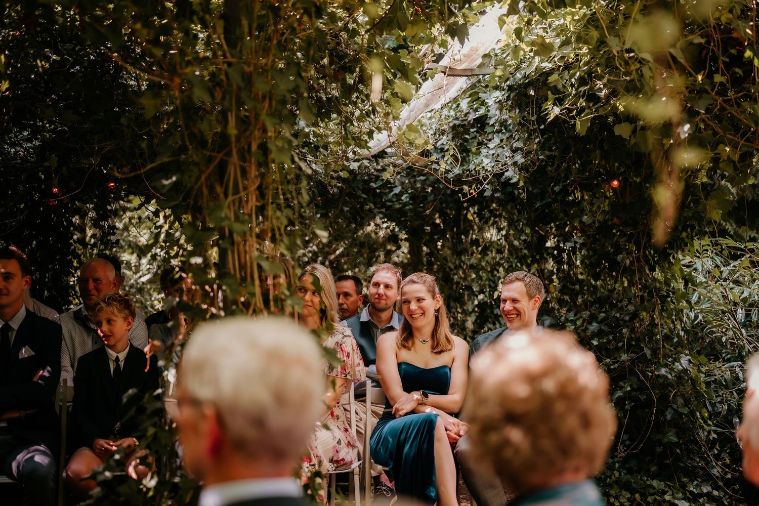 Outdoor wedding ceremony with guests seated in a garden setting surrounded by lush greenery.