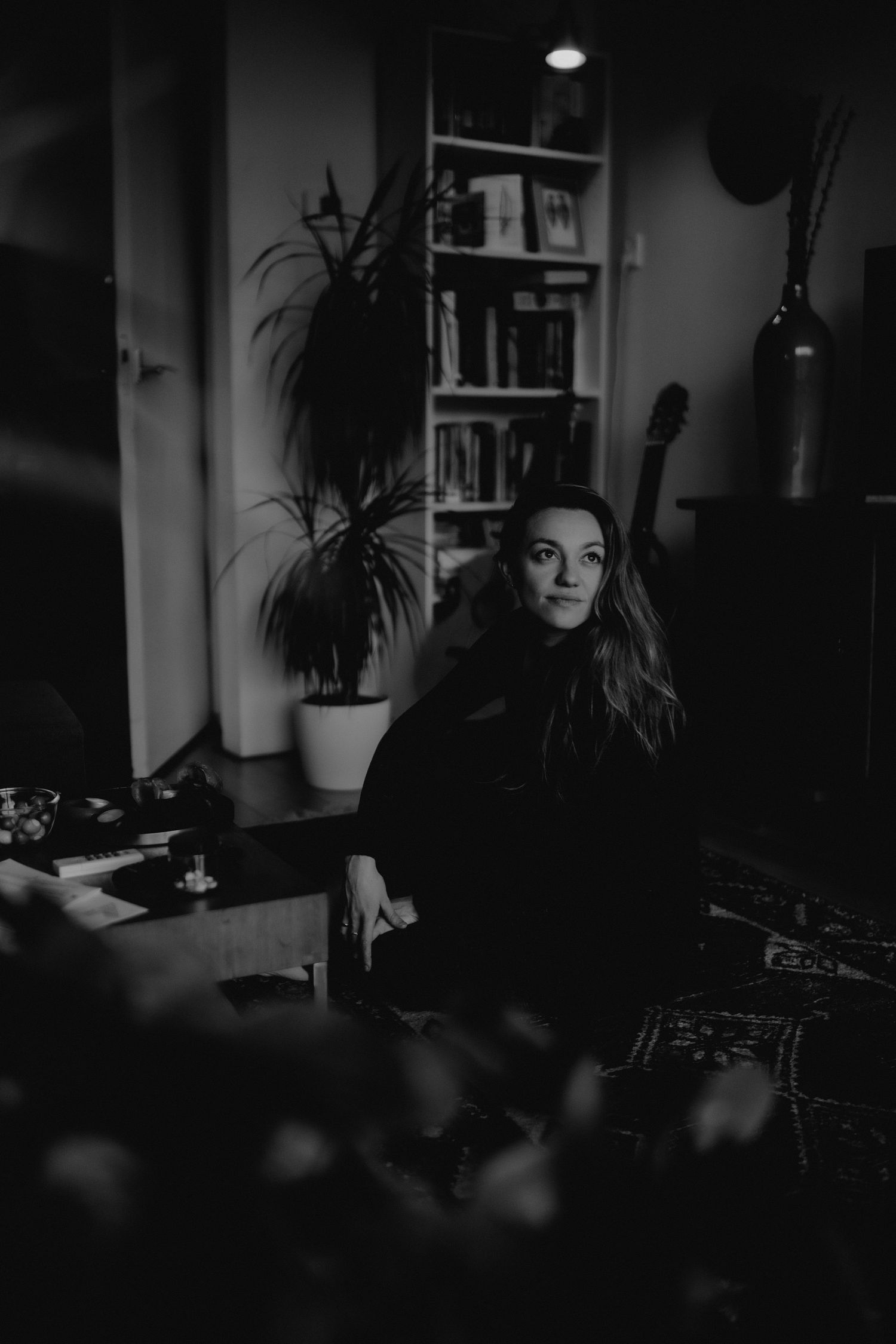 A moody black and white interior photo showing a person seated in a dark room with houseplants and bookshelves visible.