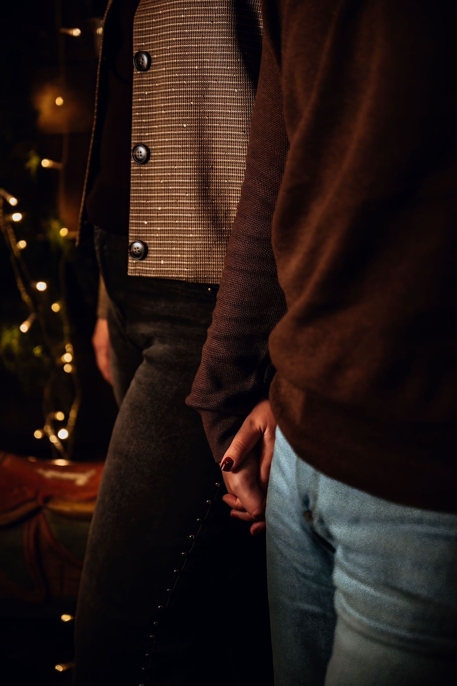 A person wearing jeans and a brown sweater stands against a dark background with twinkling lights.