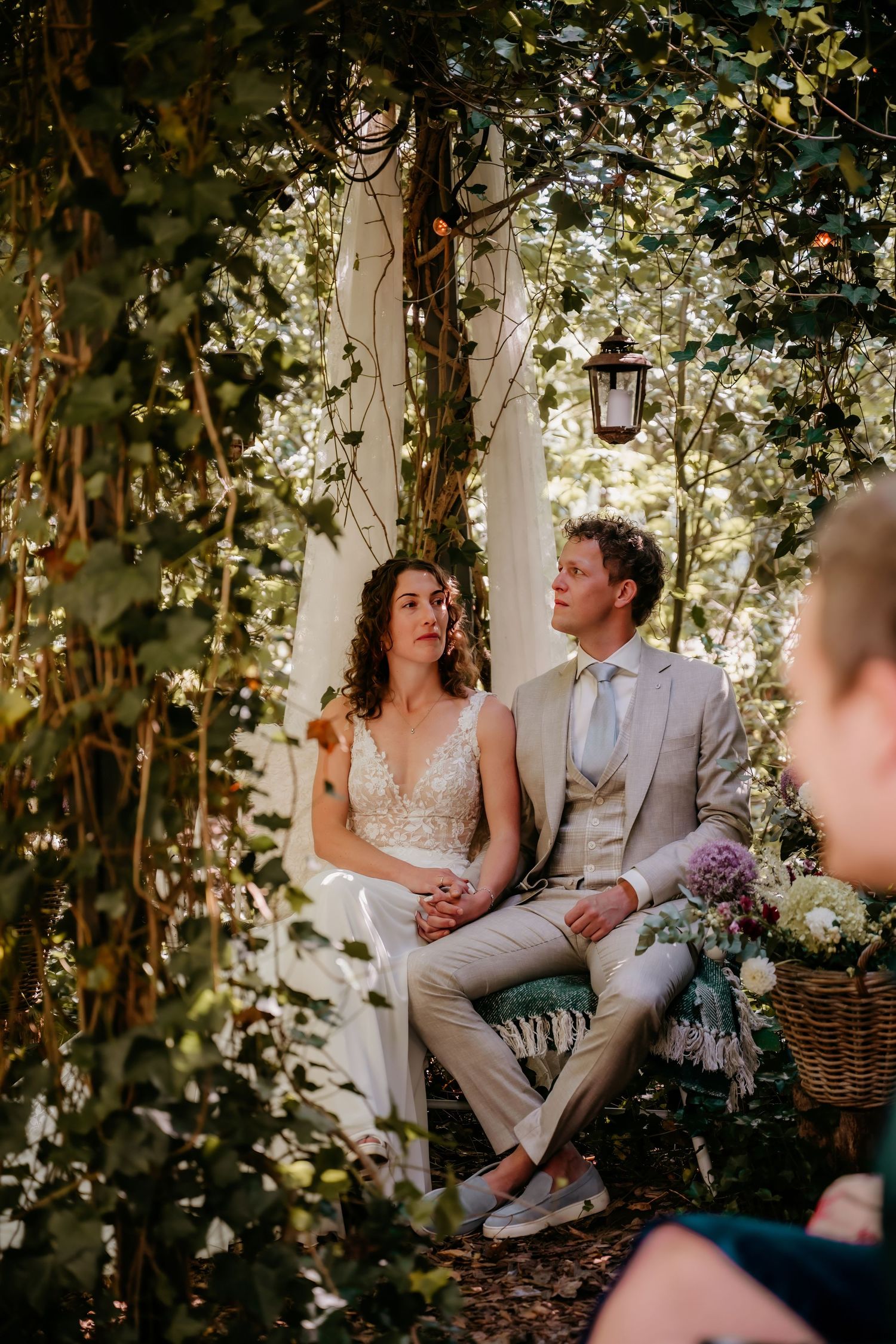 A couple shares an intimate moment on a bench surrounded by lush greenery and hanging lights at an outdoor wedding.