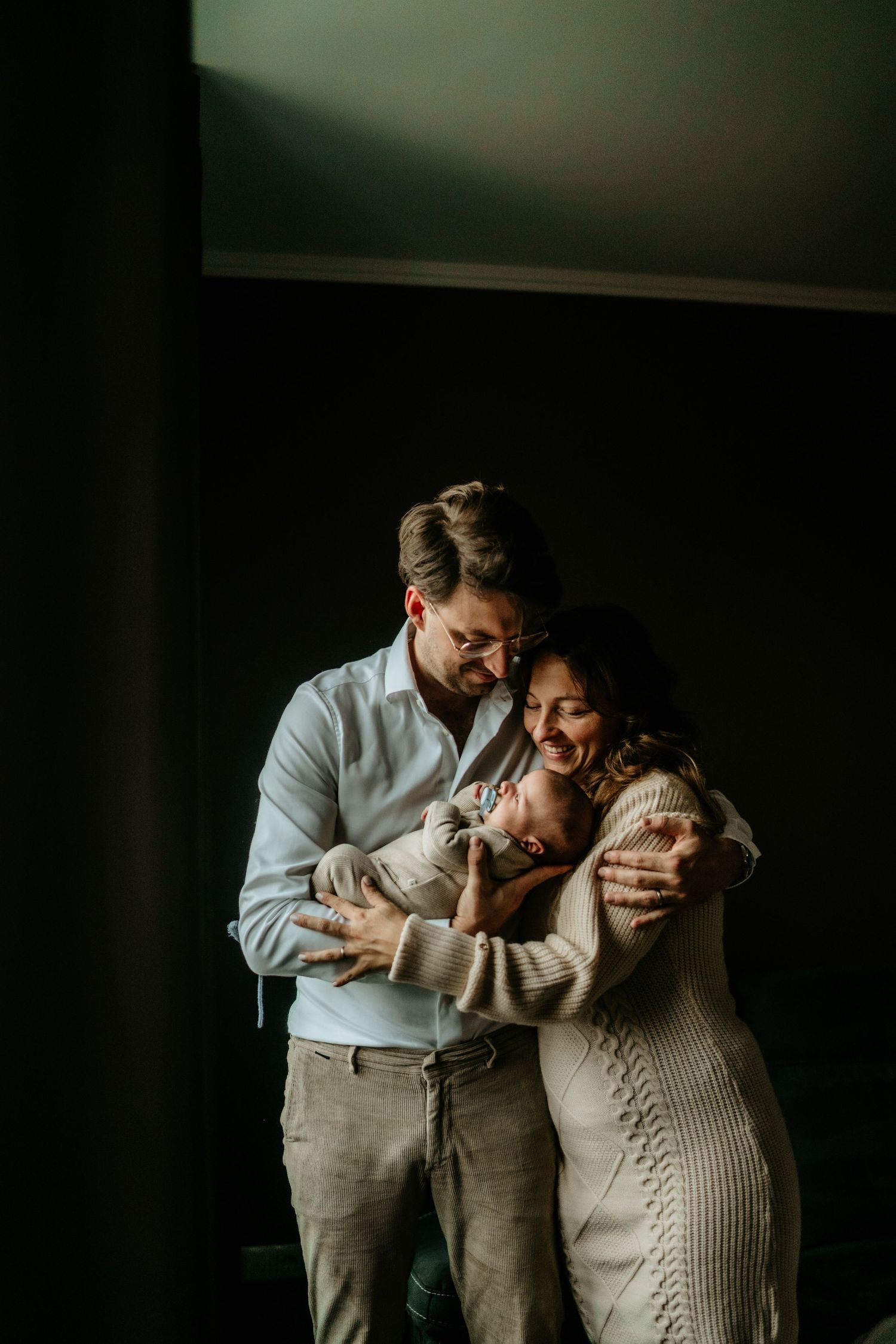 Intimate portrait of couple holding newborn baby in moody indoor lighting.