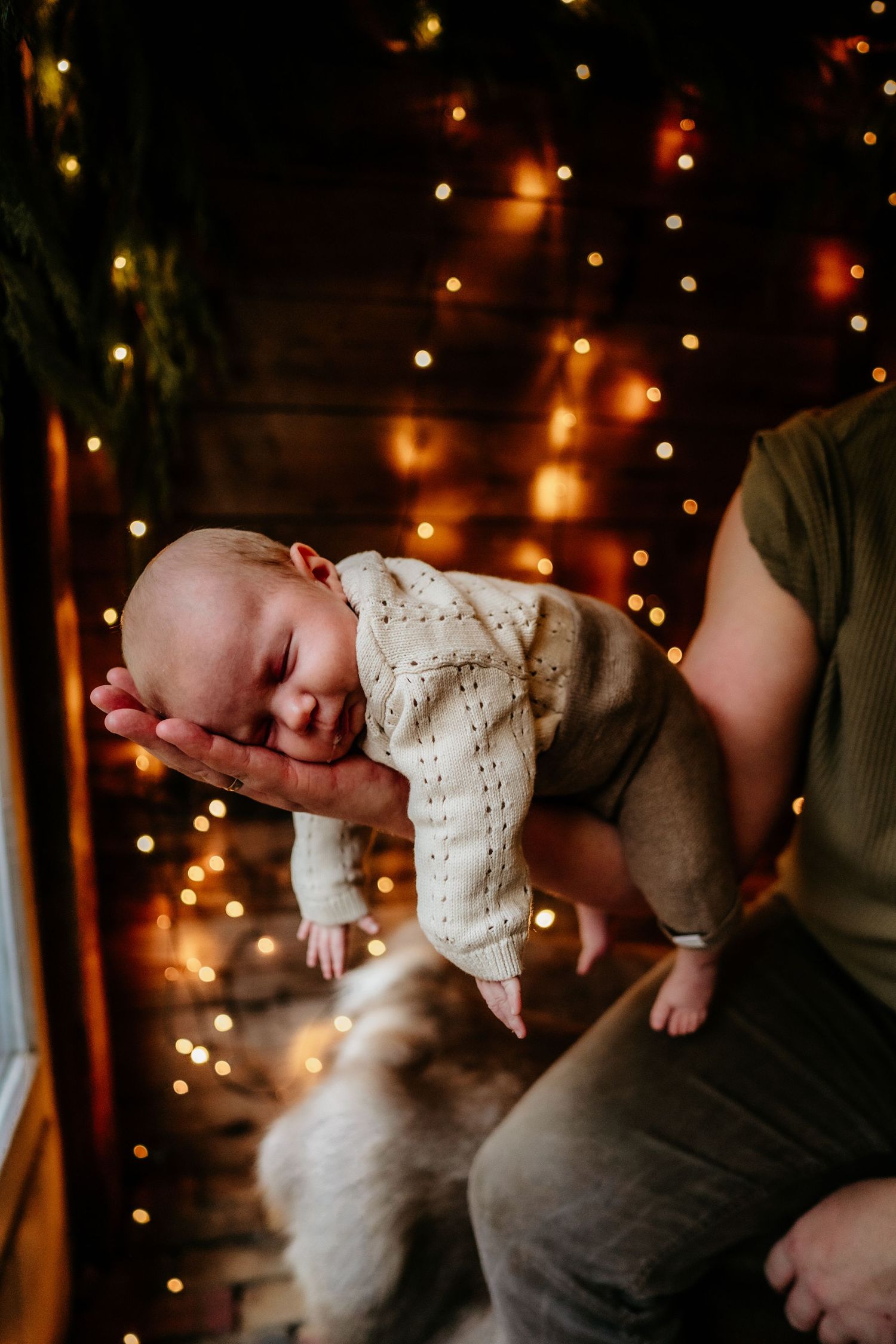 Sleeping newborn baby in knit outfit cradled in hands against glowing holiday lights background.
