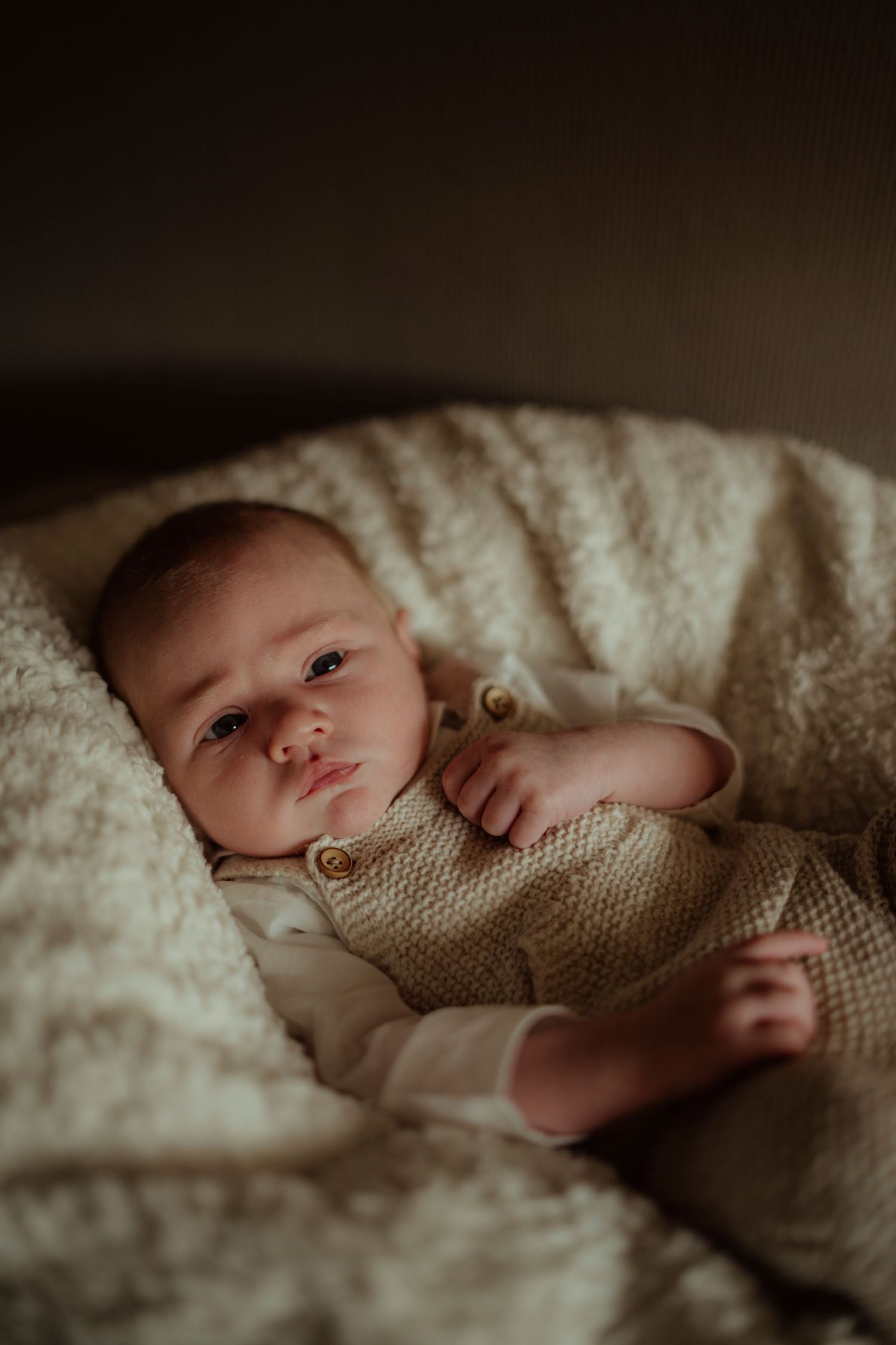 A baby in a knitted onesie lies snuggled in white blankets with soft lighting.