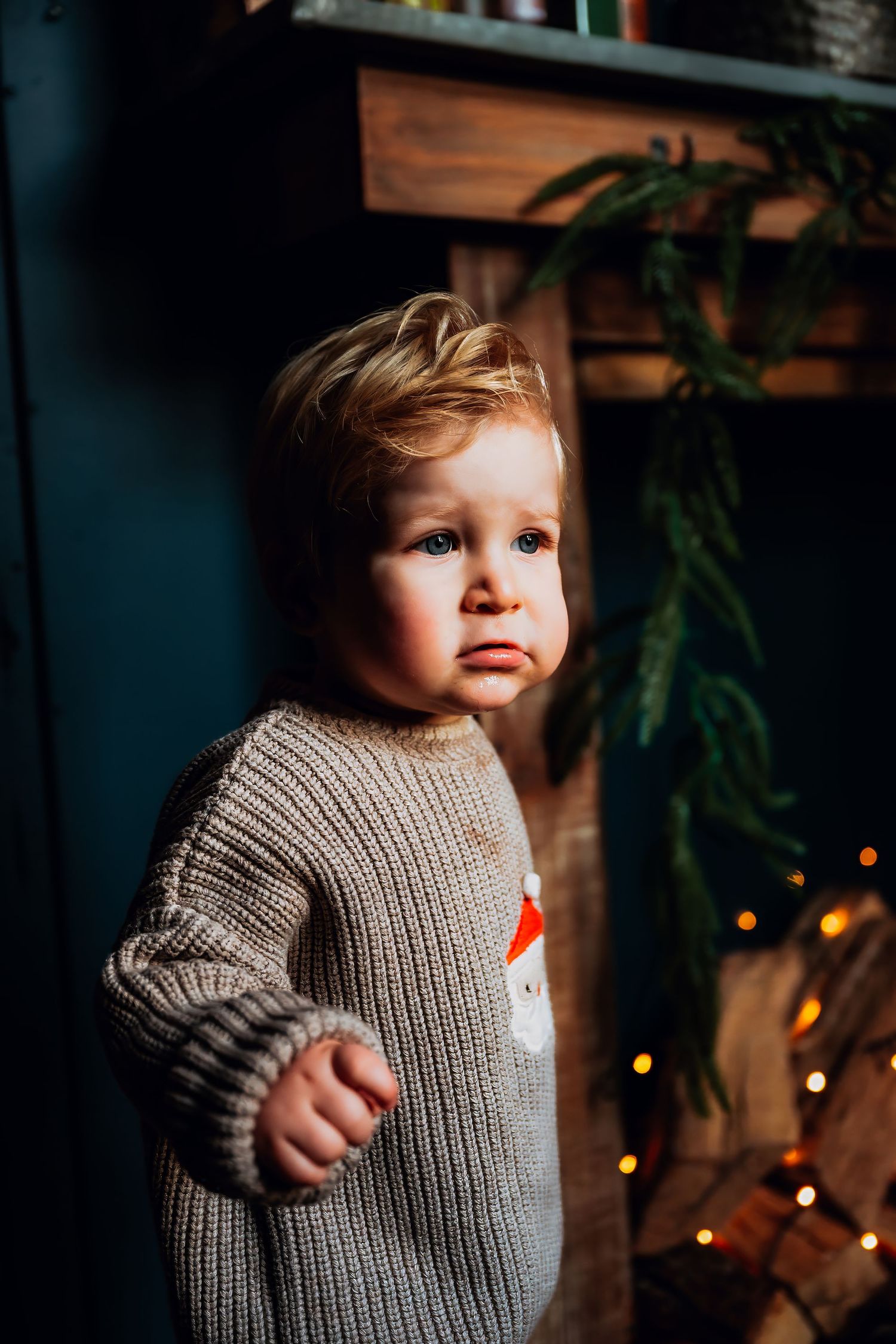 Child wearing a gray knit sweater stands against a dark background with twinkling lights.