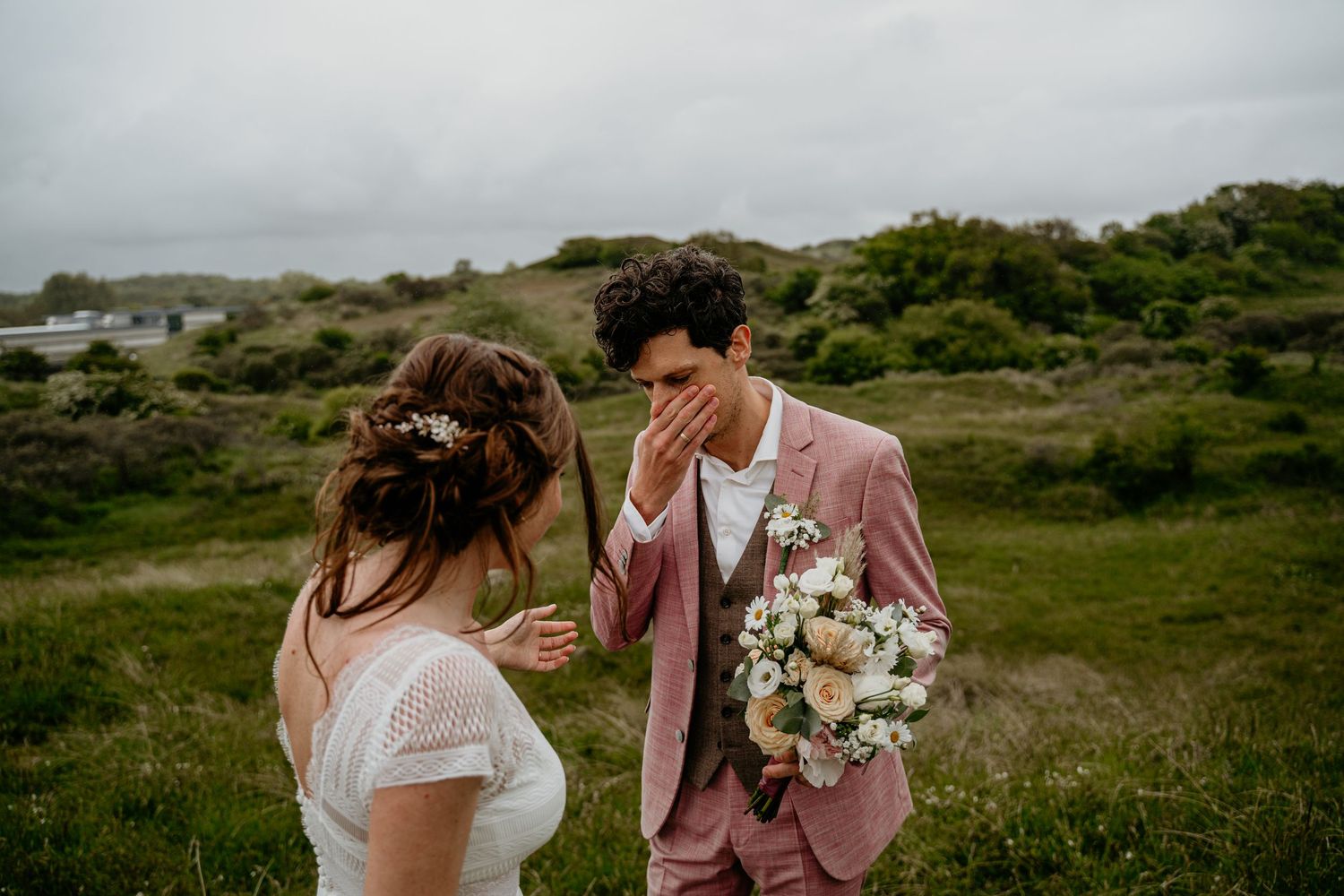 Couple in wedding attire walking together through a scenic coastal landscape with green hills.