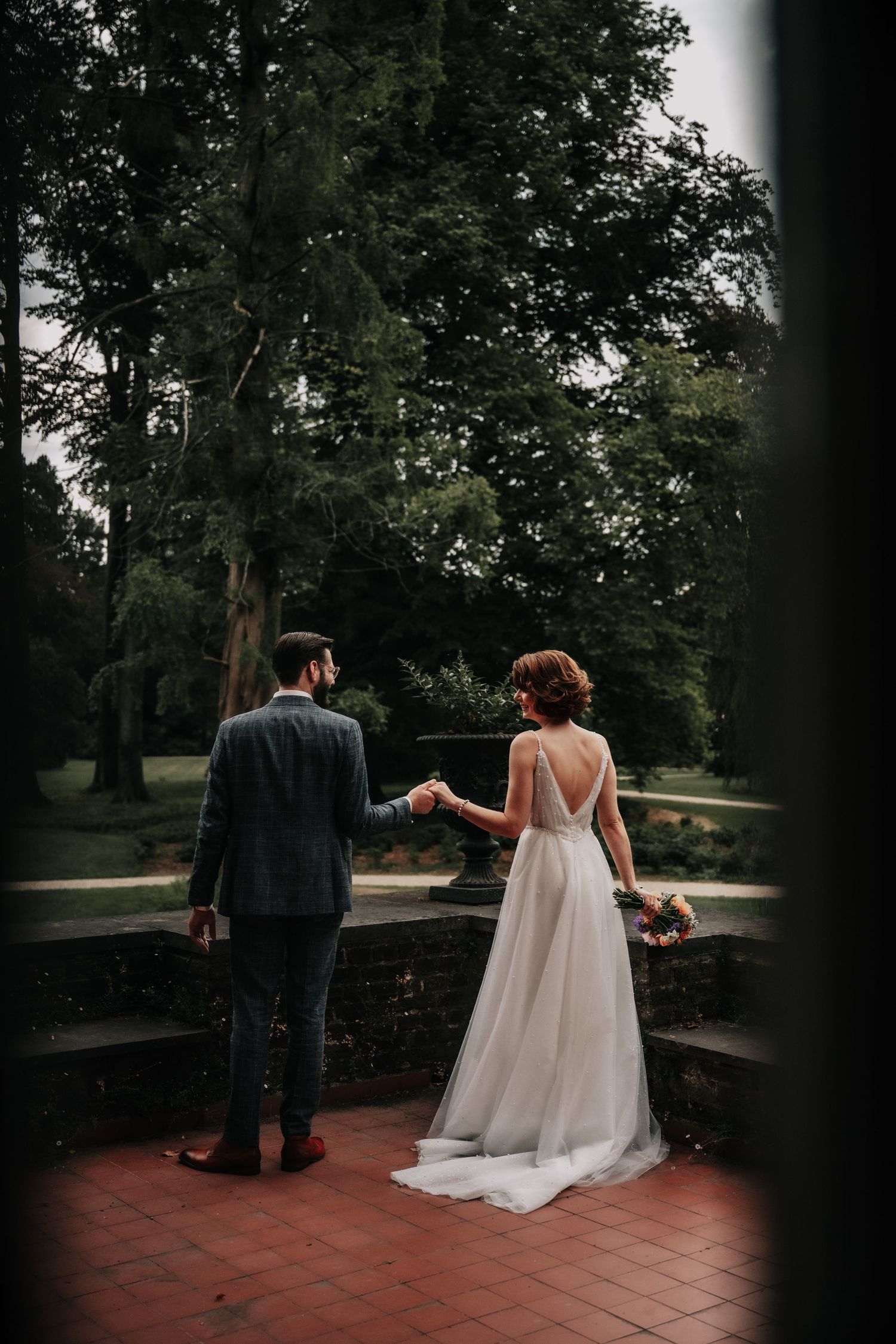 A couple walks along a brick terrace surrounded by lush trees on a cloudy day.