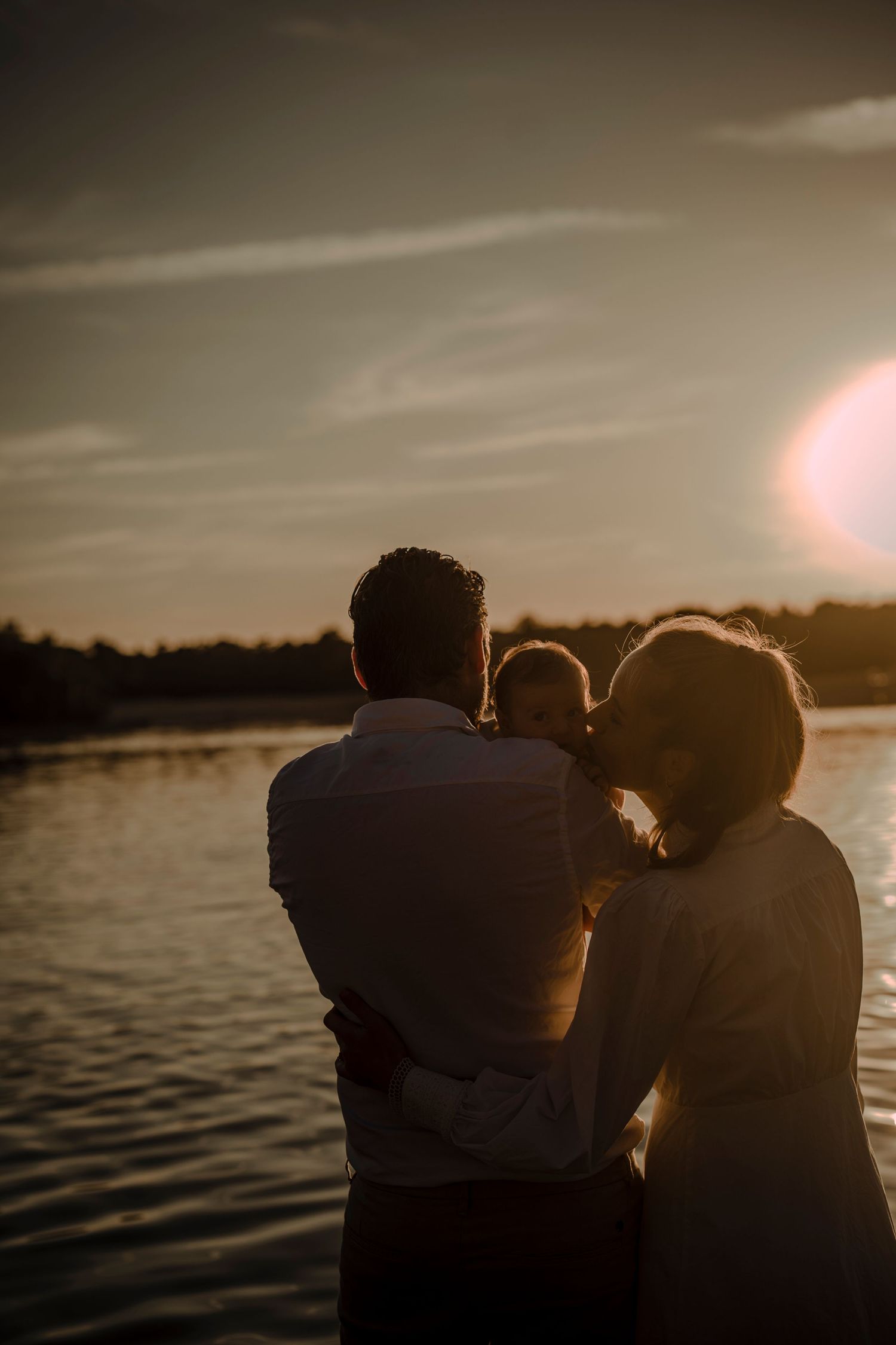A couple stands silhouetted against a warm sunset over a serene lake, creating a romantic backlit scene.
