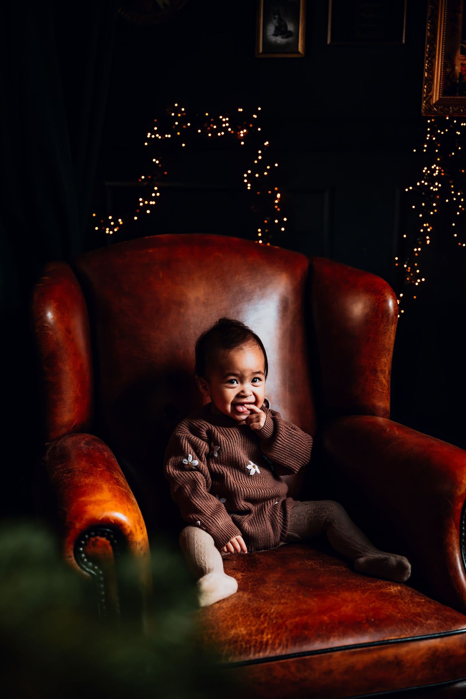 A happy toddler in brown sweater sits in an oversized leather armchair with holiday decorations behind.