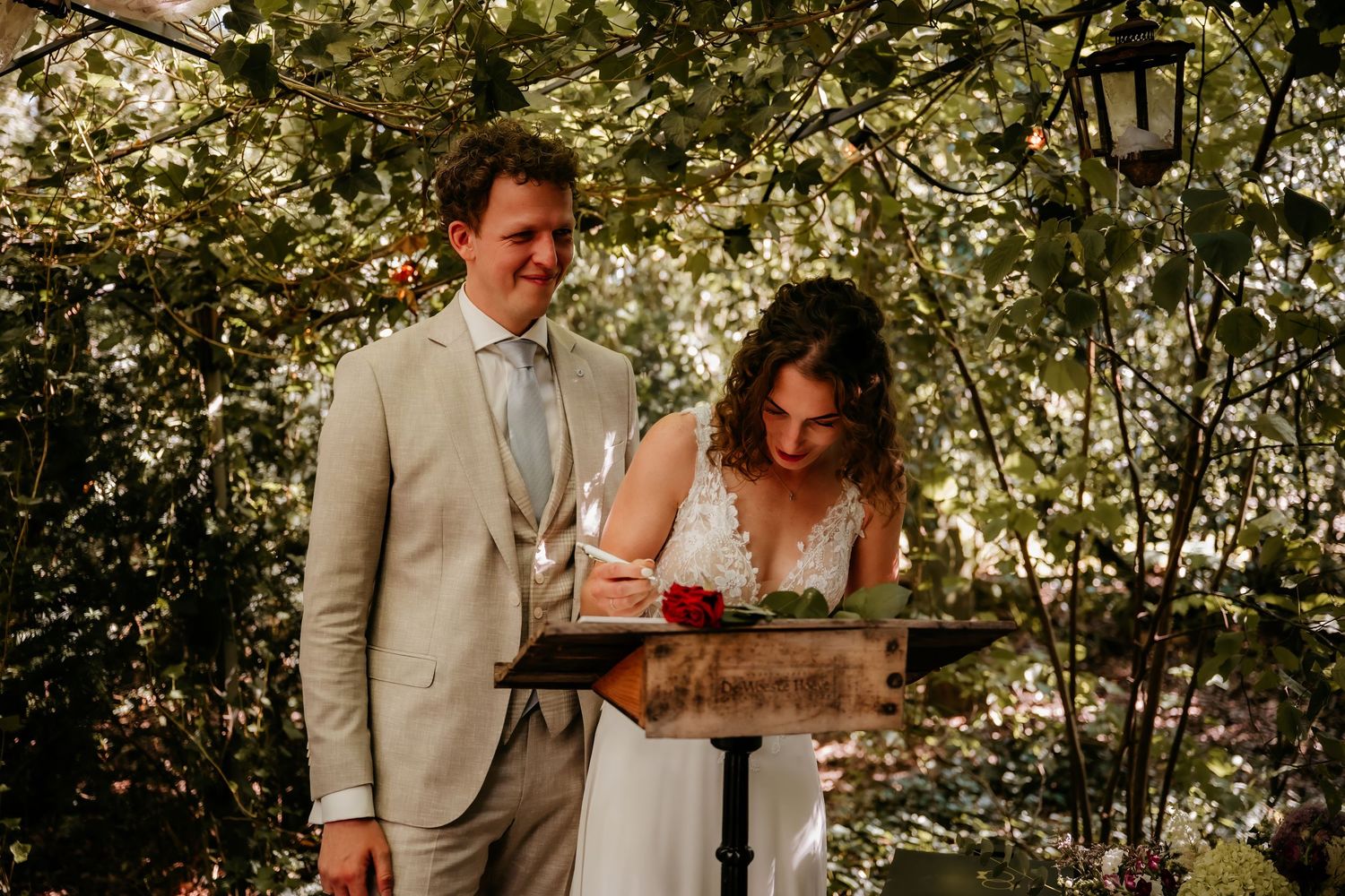 Wedding ceremony taking place under lush green trees in an outdoor garden setting.