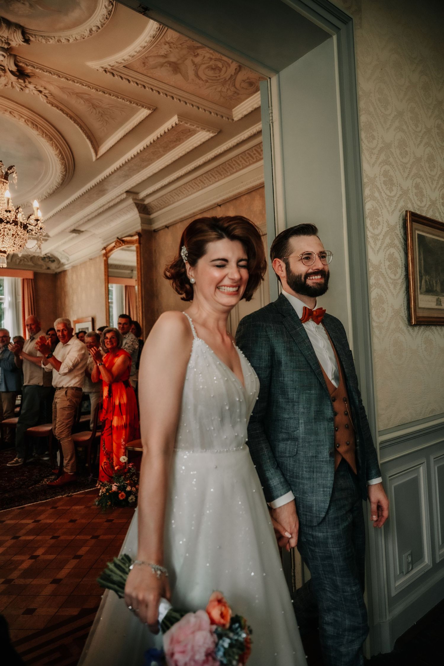 Newlyweds walk together through an elegant ballroom with ornate moldings and blue-grey walls.