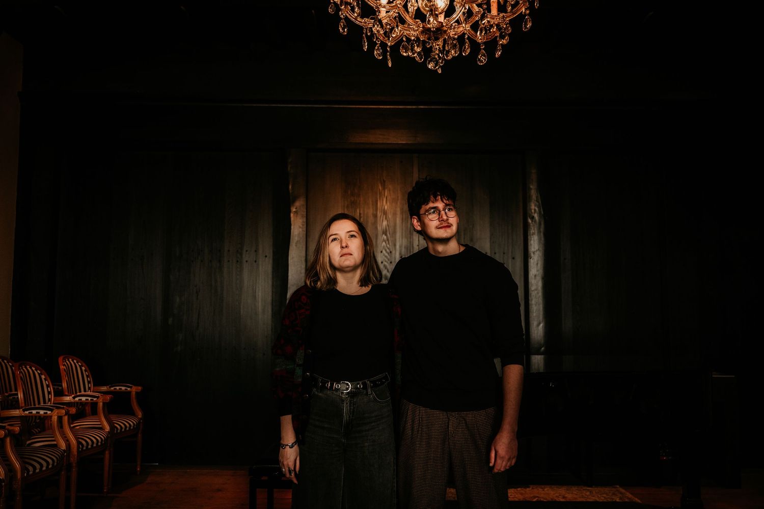 Two people stand together in a dark moody room with wooden walls and an ornate chandelier overhead.