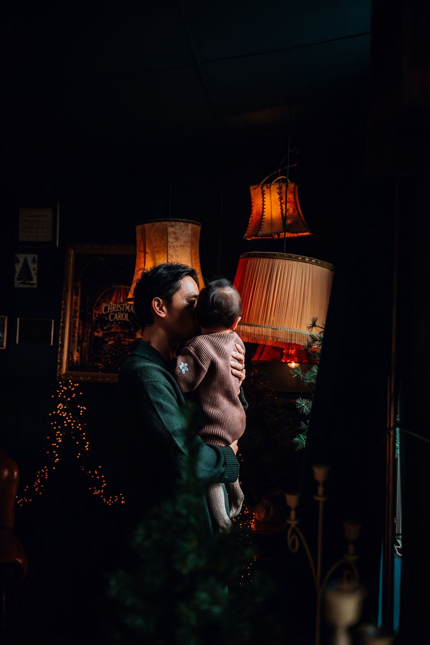 A romantic couple embraces in a dimly lit room with warm glowing lamps and twinkling holiday lights in the background.