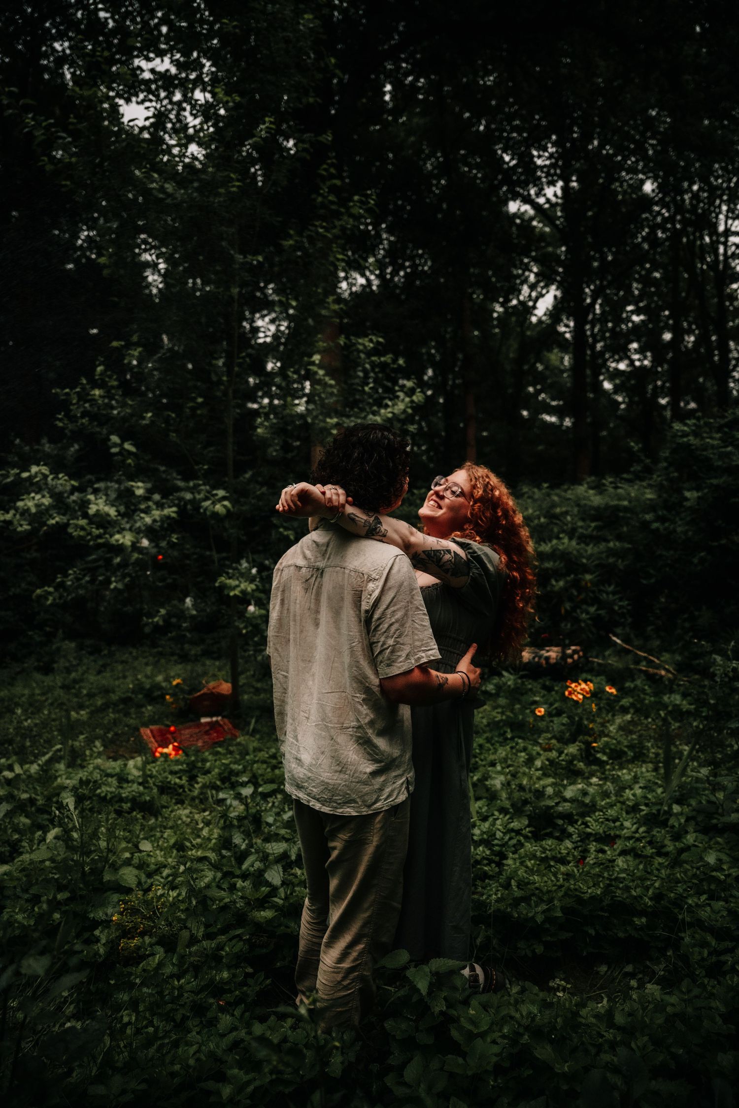 Two figures stand close together in a moody garden setting with dark foliage in the background.