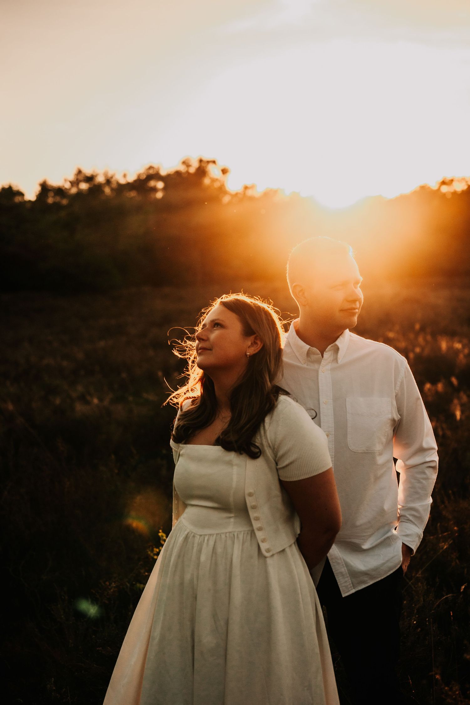 A couple in white dress attire poses together during a romantic sunset photoshoot in a golden field.
