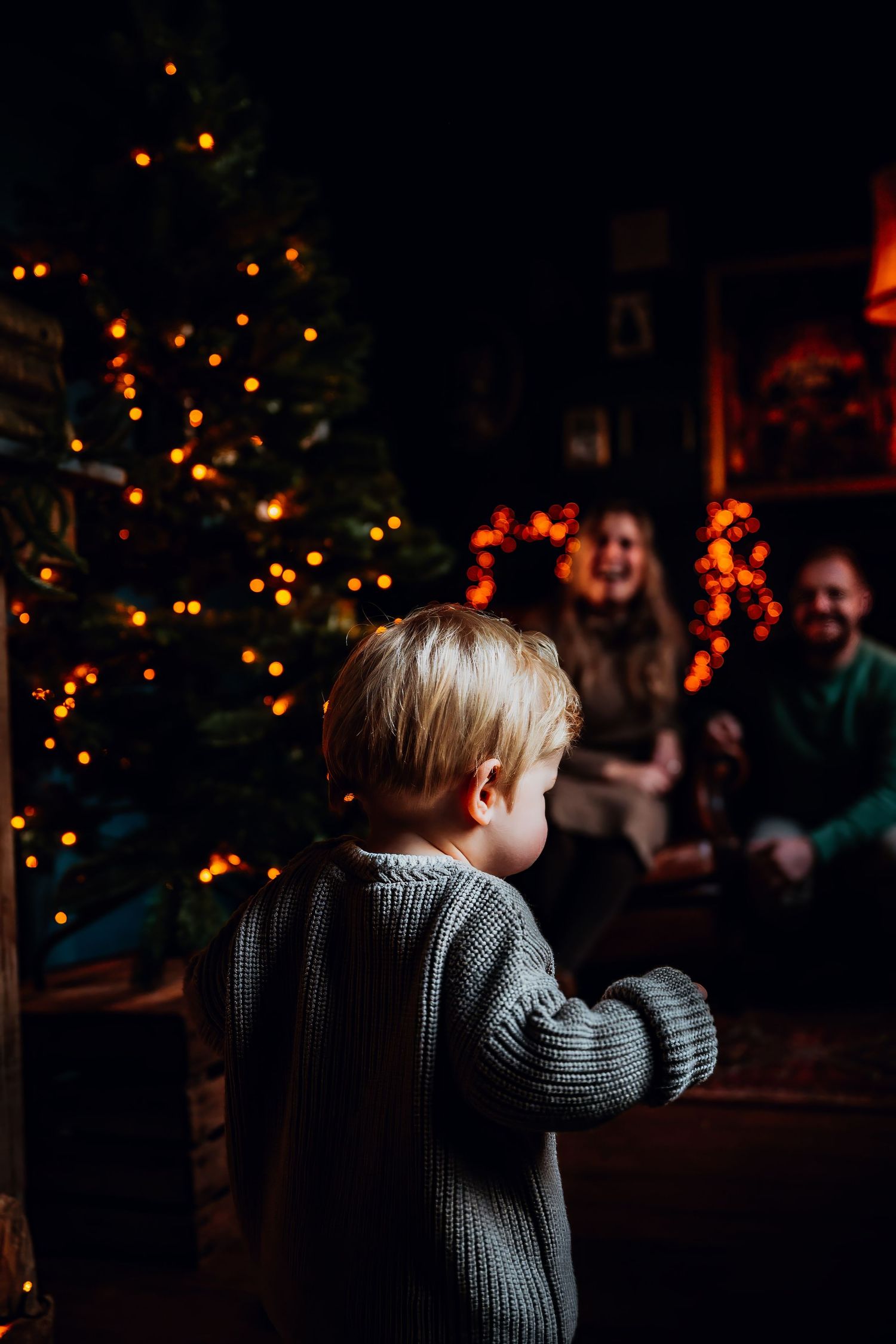 Child in gray sweater stands against dark background with glowing holiday lights creating a warm, moody atmosphere.