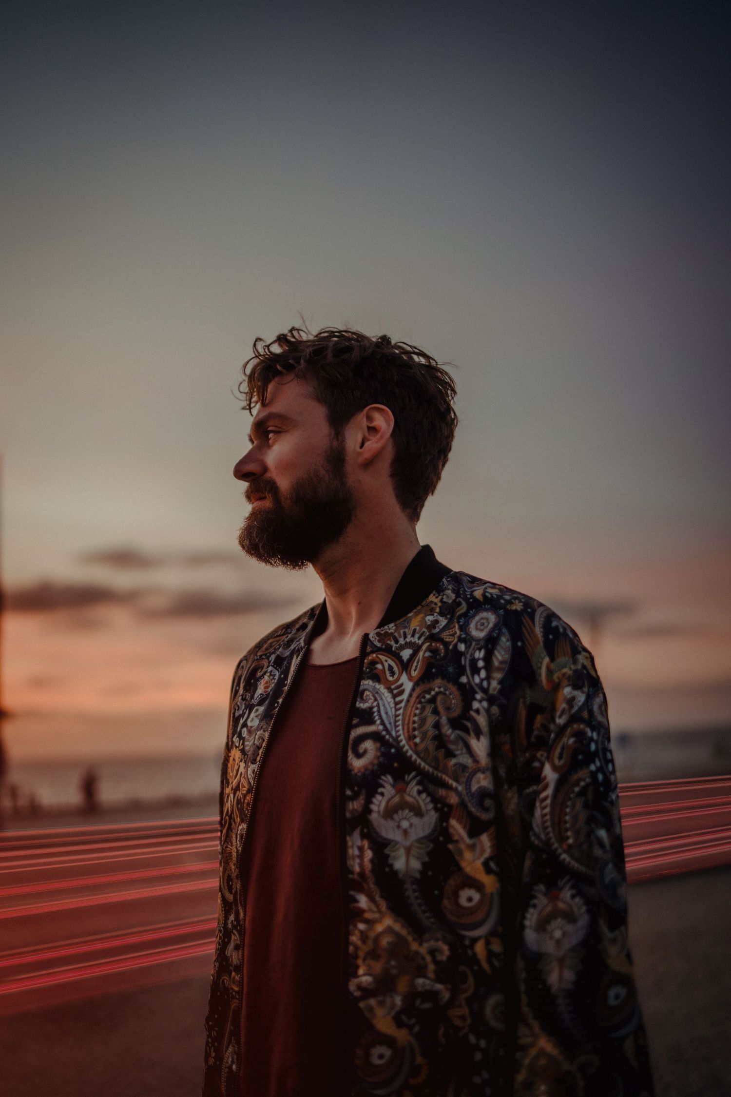Profile silhouette of person wearing patterned jacket against sunset sky with light trails in background.
