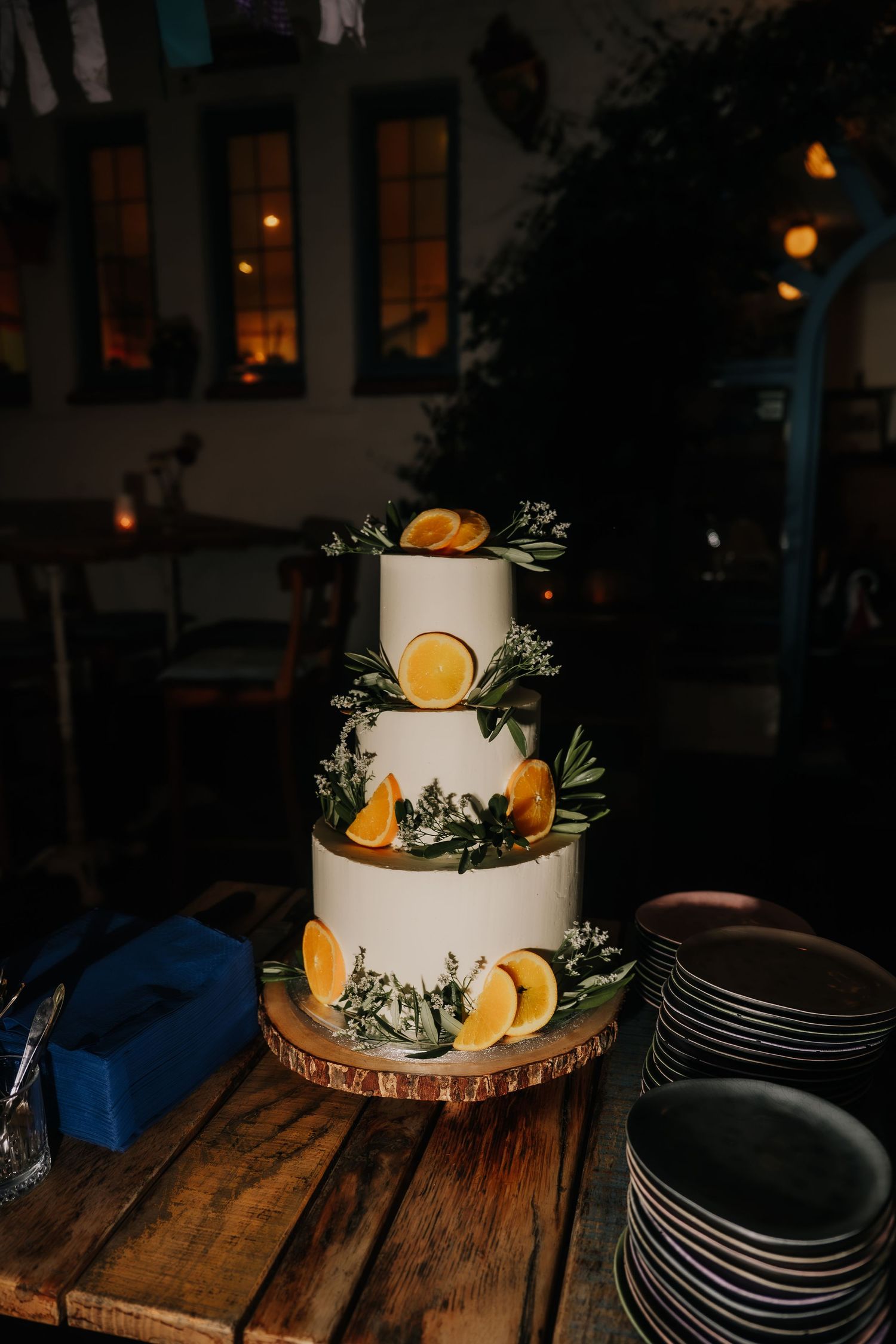 Elegant three-tier white wedding cake decorated with fresh orange slices and greenery on a rustic wood stand.