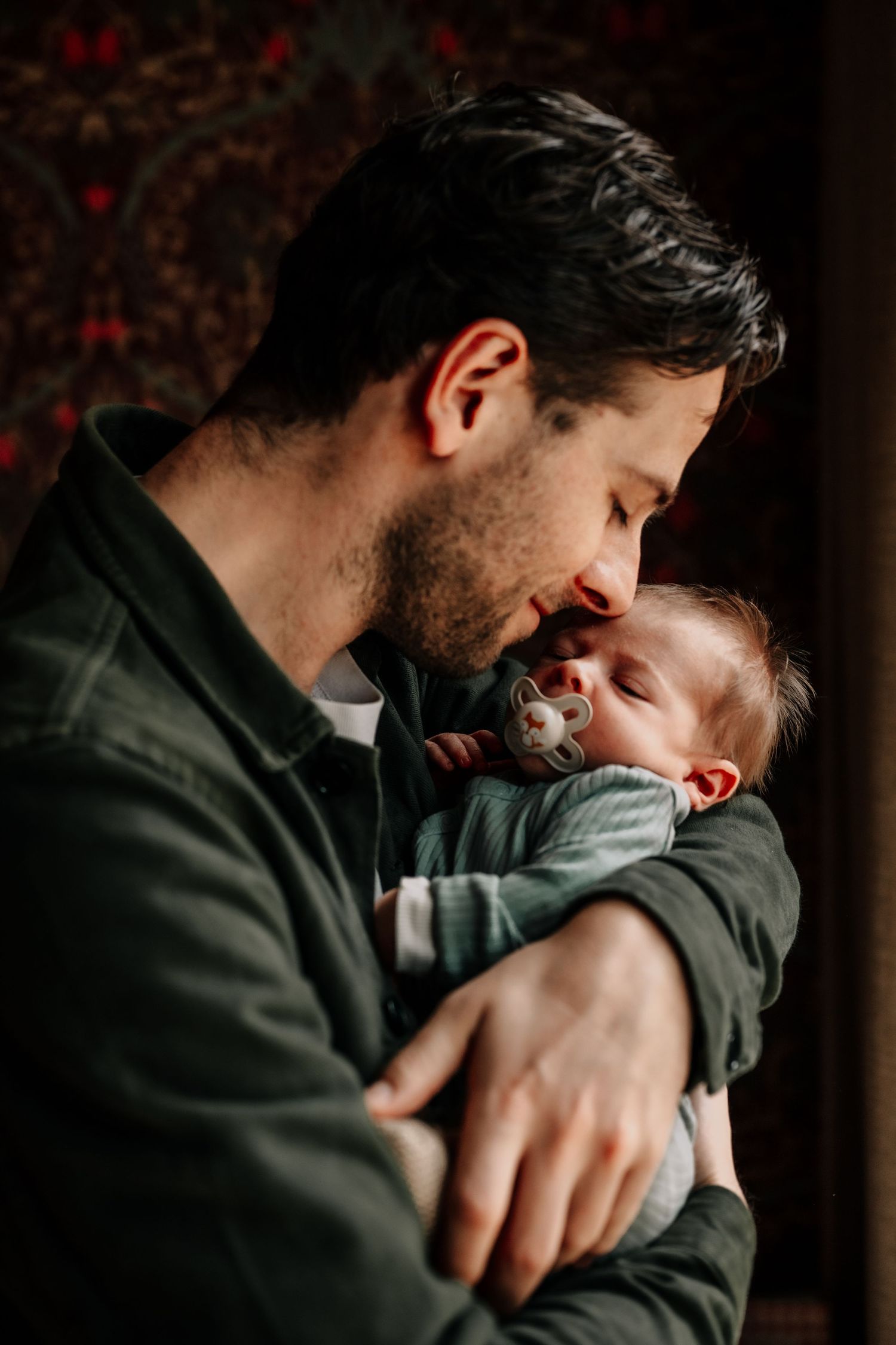 Close-up of a gentle moment with a newborn against dark patterned wallpaper.
