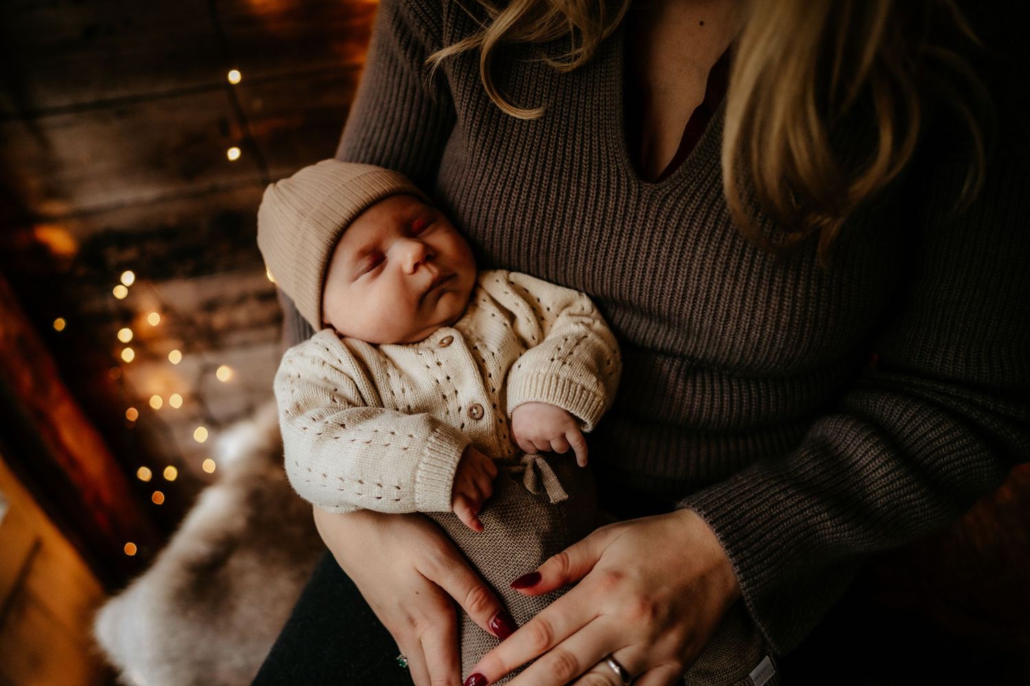 Infant in winter attire resting in caregiver's arms with holiday lights in background.