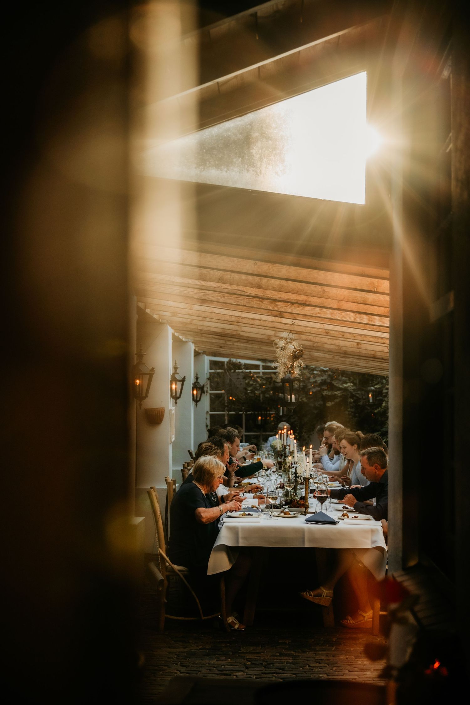 Warm evening light streams into a rustic reception venue where wedding guests dine at long tables.
