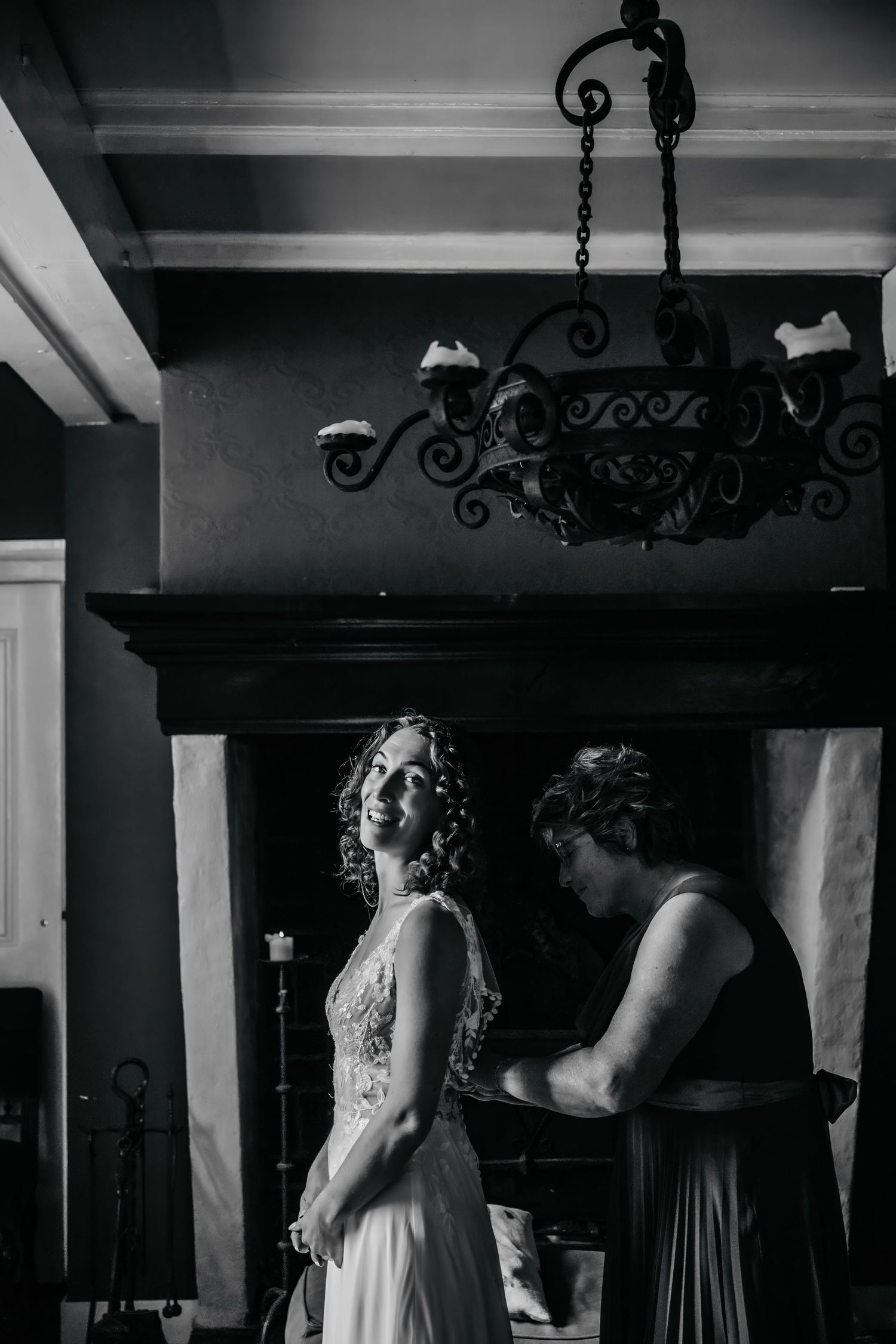 Black and white photo of a wedding dress preparation scene under an ornate iron chandelier.