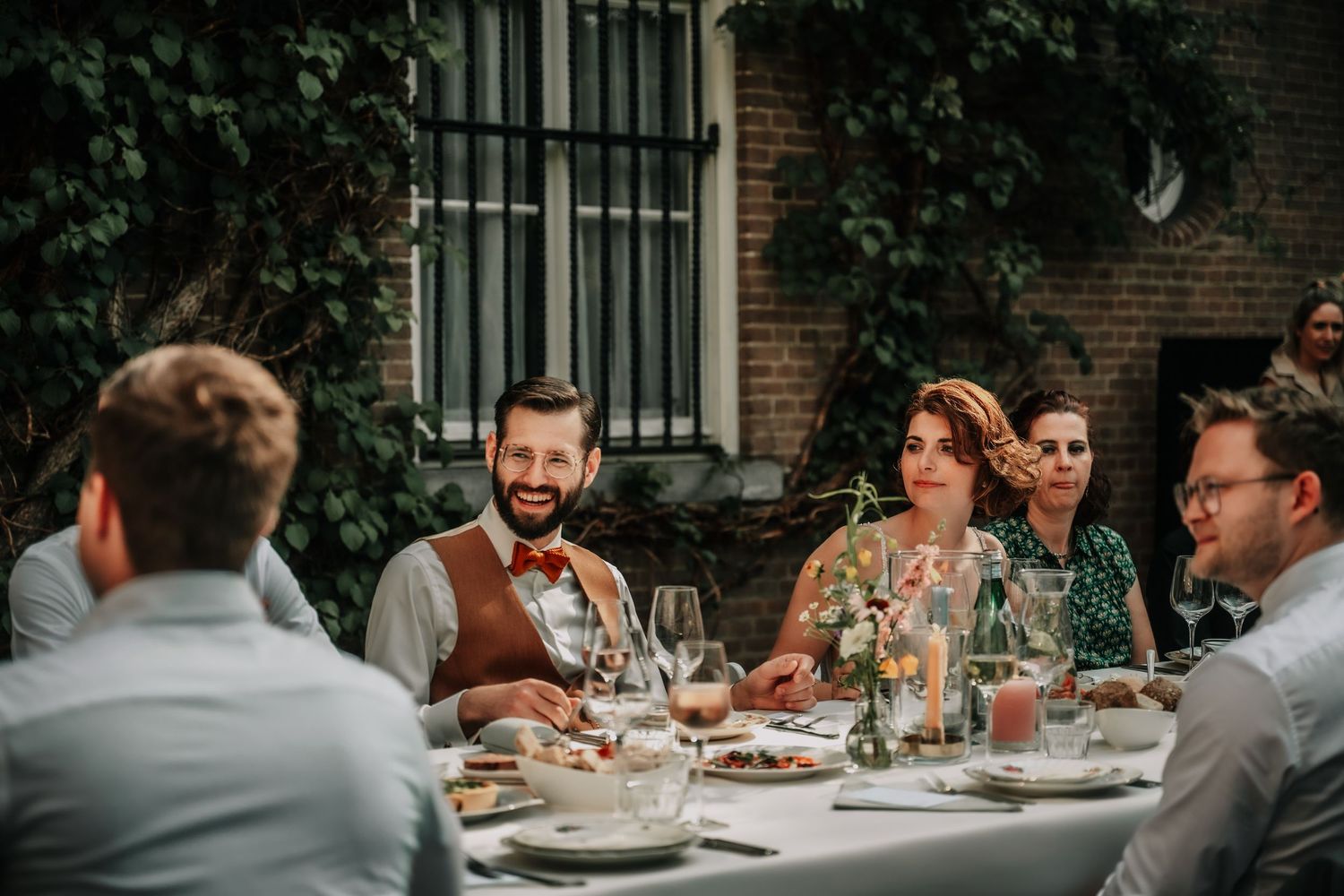 Outdoor wedding dinner setting with brick wall and greenery backdrop.