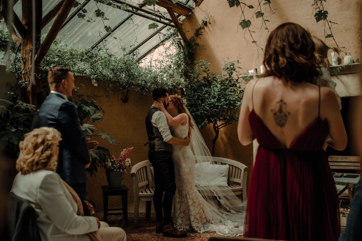 An intimate indoor wedding ceremony under a greenhouse-style roof with greenery and decorations.