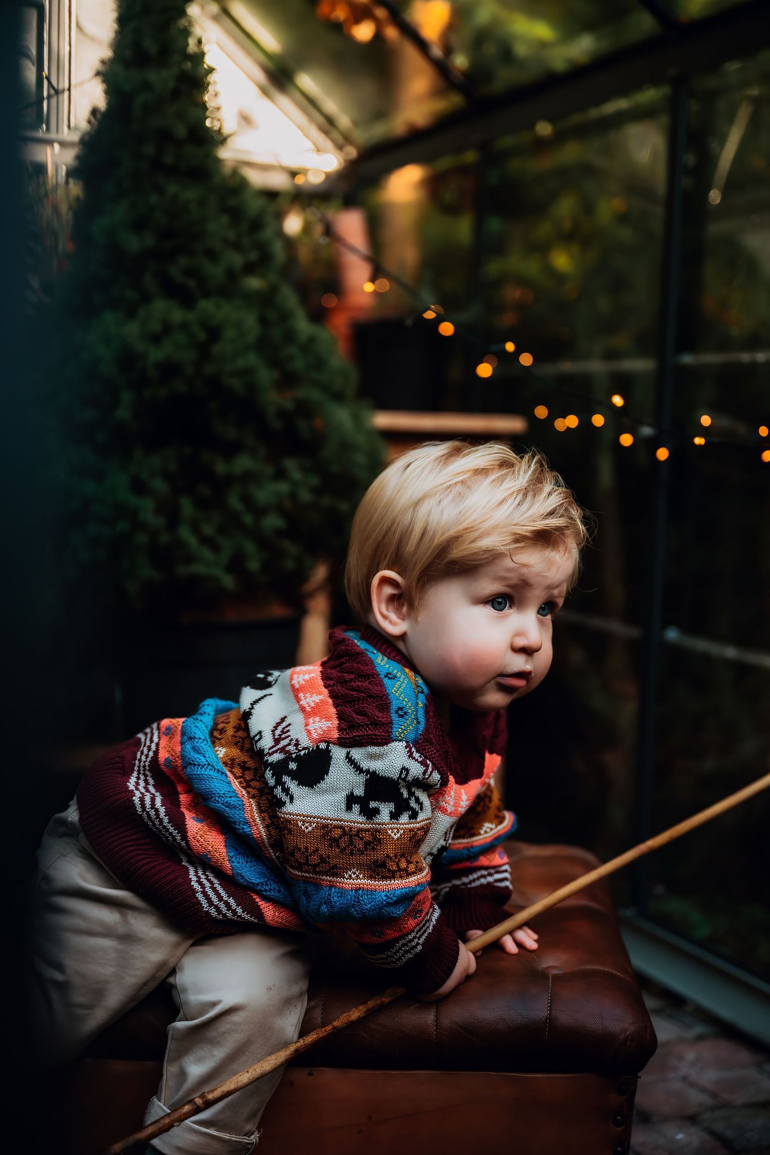 A child leans over a dark surface while holding a wooden stick in a dimly lit room with twinkling lights in background.