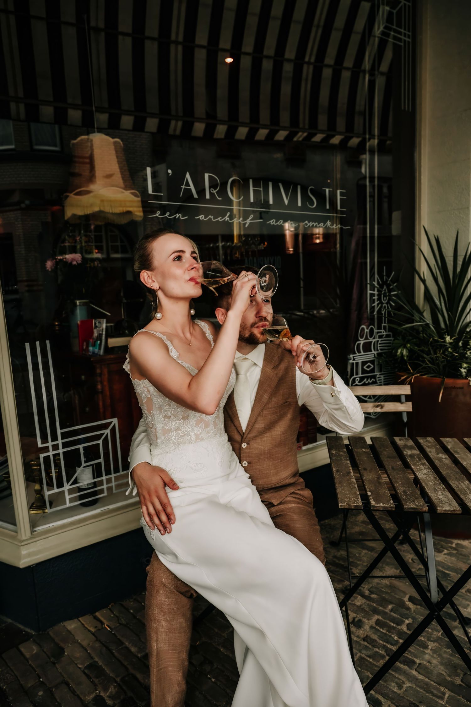 Bride and groom sipping champagne outside L'Archiviste café, wearing elegant wedding attire on a charming brick patio.