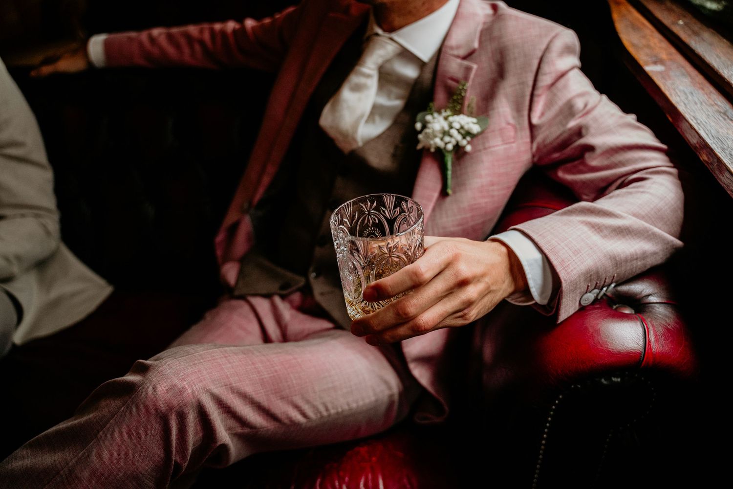 Person wearing a pink suit holds a glass while sitting in moody lighting.