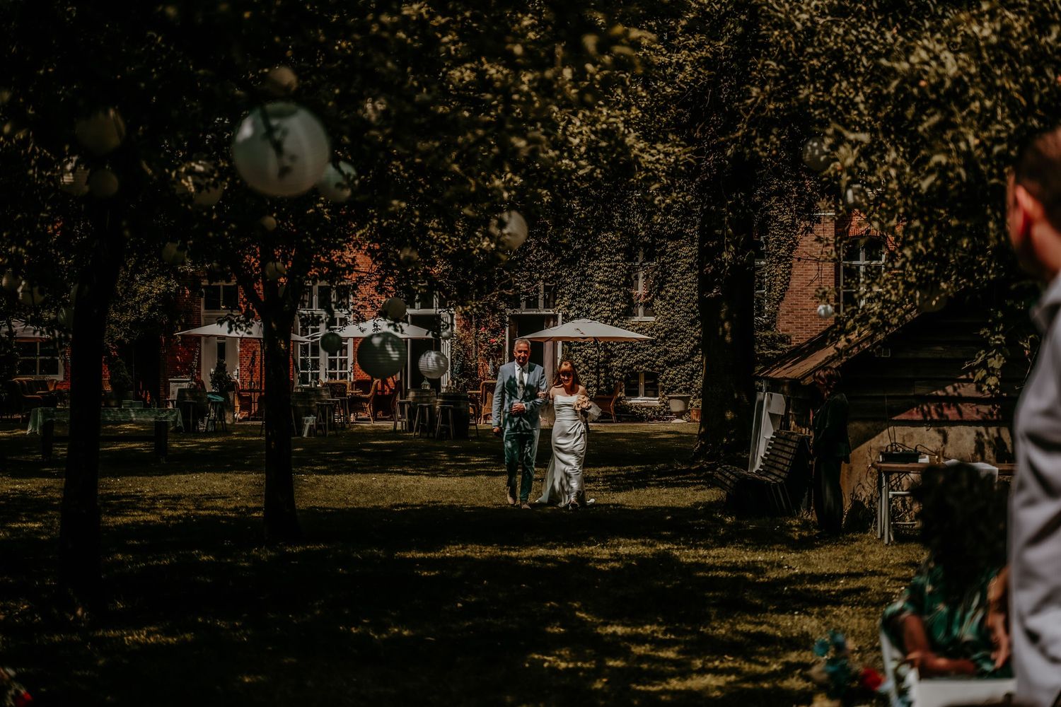 People enjoying a sunny day in a park with trees and umbrellas casting shadows on the grass.