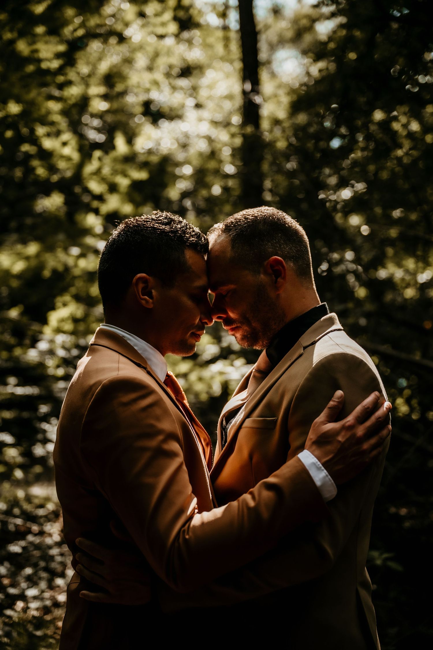 Two people in tan suits embrace intimately in a sunlit forest setting with bokeh effect in the background.