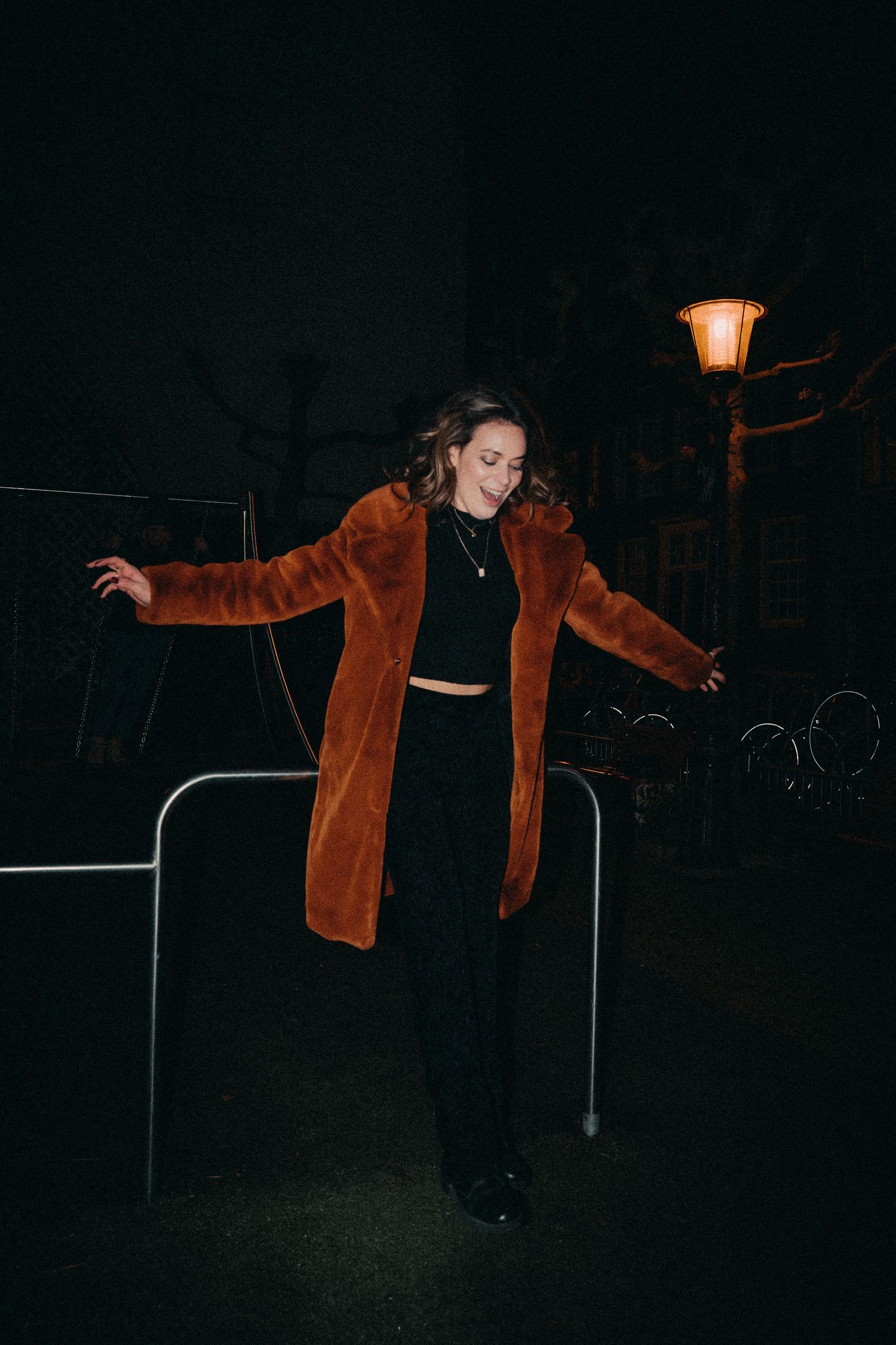 A person in a brown jacket balances on metal railings at night with a glowing street lamp in the background.