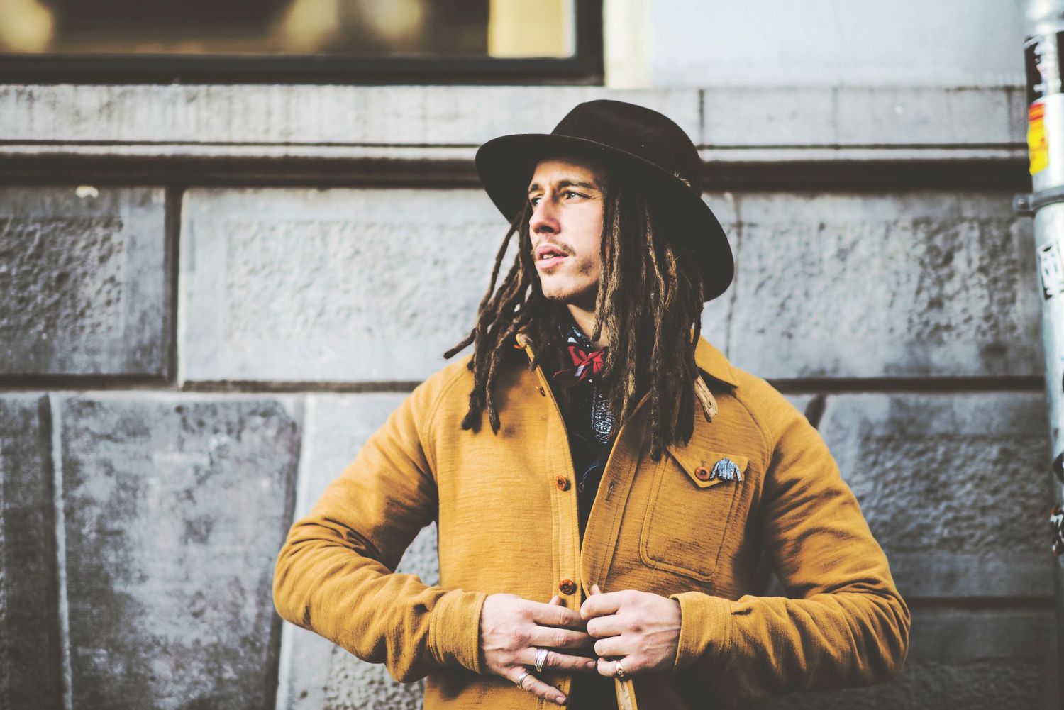 Person wearing a yellow jacket and black hat stands against stone wall in urban setting.