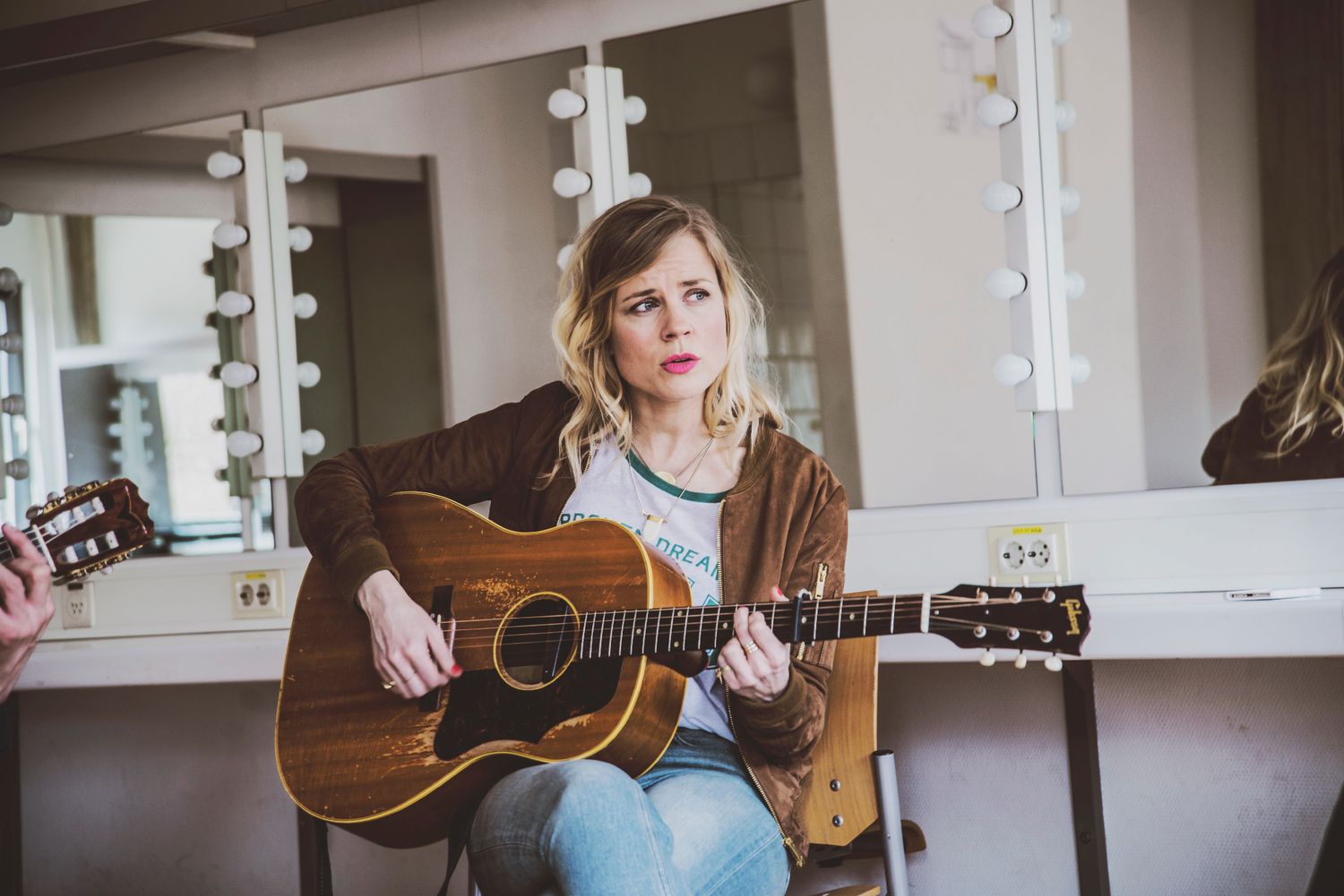 A musician performs on an acoustic guitar in a bright interior space with multiple mirrored reflections.
