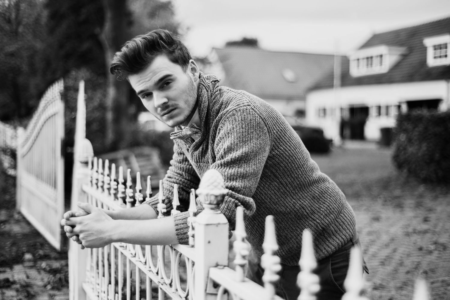 Black and white photo of someone leaning on a white picket fence with suburban houses in the background.