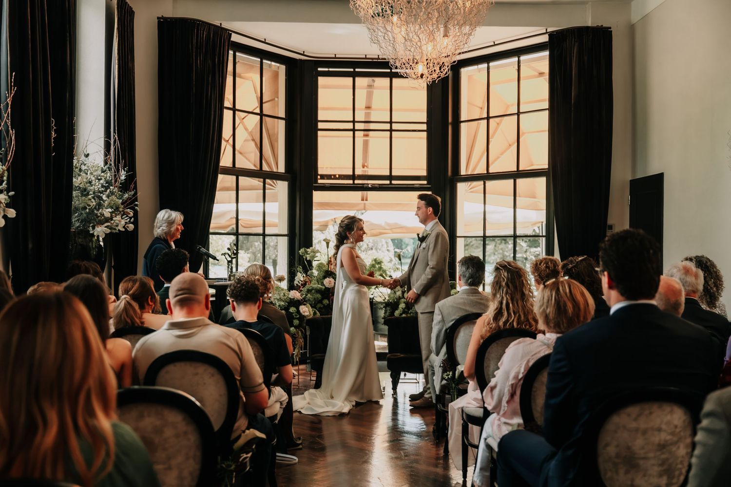 Elegant indoor wedding ceremony with bride and groom at altar, guests seated, chandelier overhead, large bay windows backdrop.