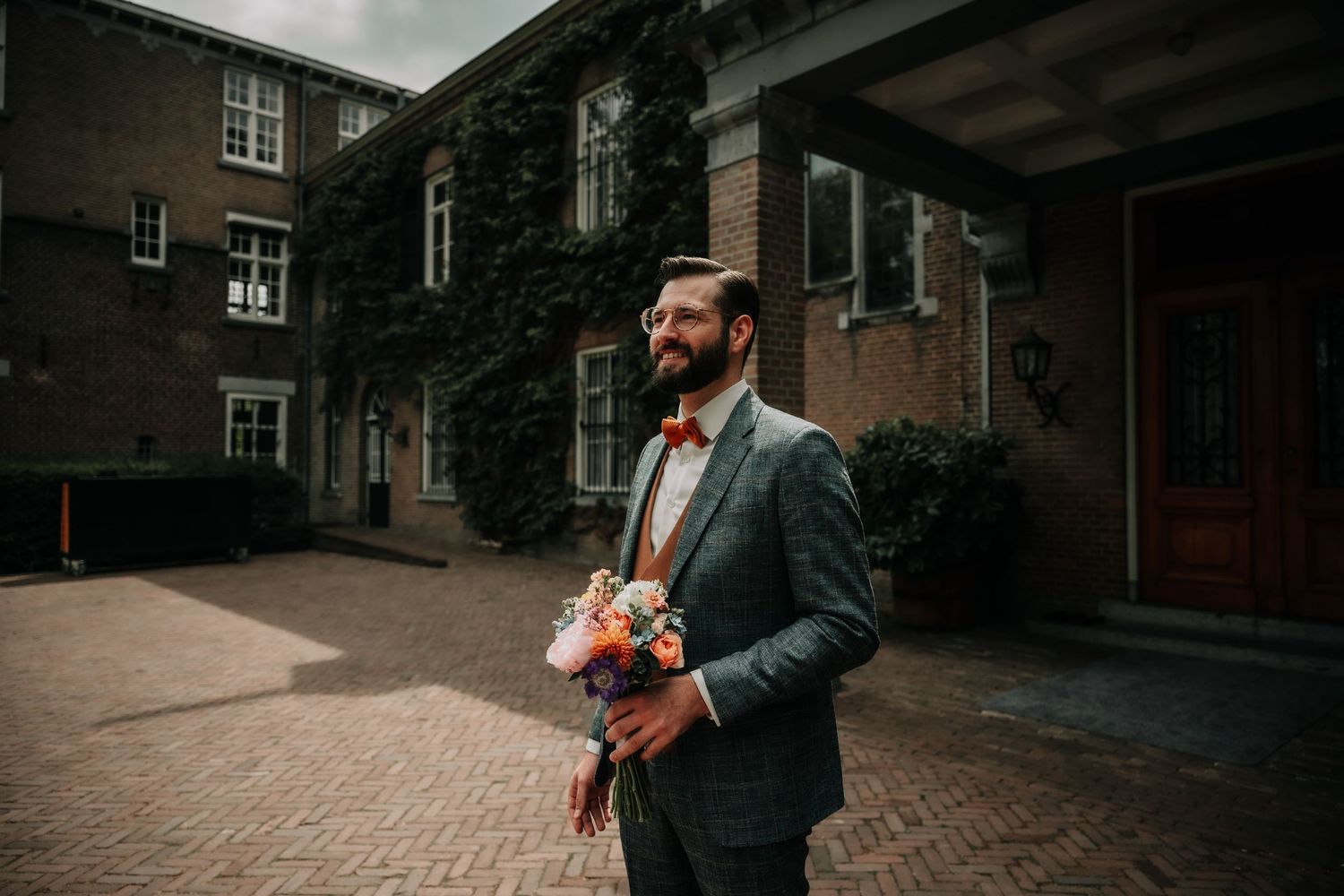 A person in a suit and tie stands in front of a brick building on a cloudy day.