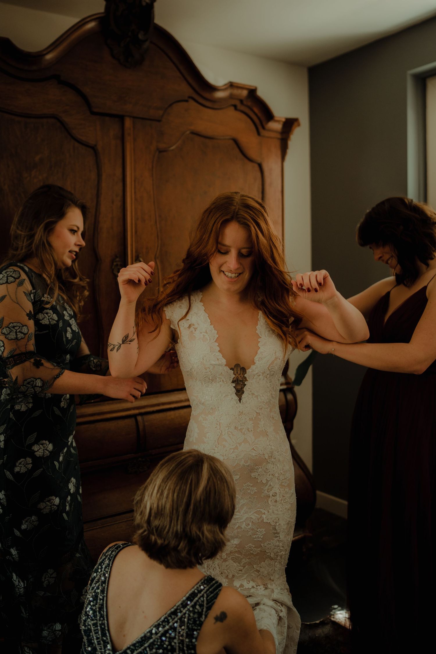 A bride getting help putting on her fitted lace wedding dress in front of an antique wooden armoire.