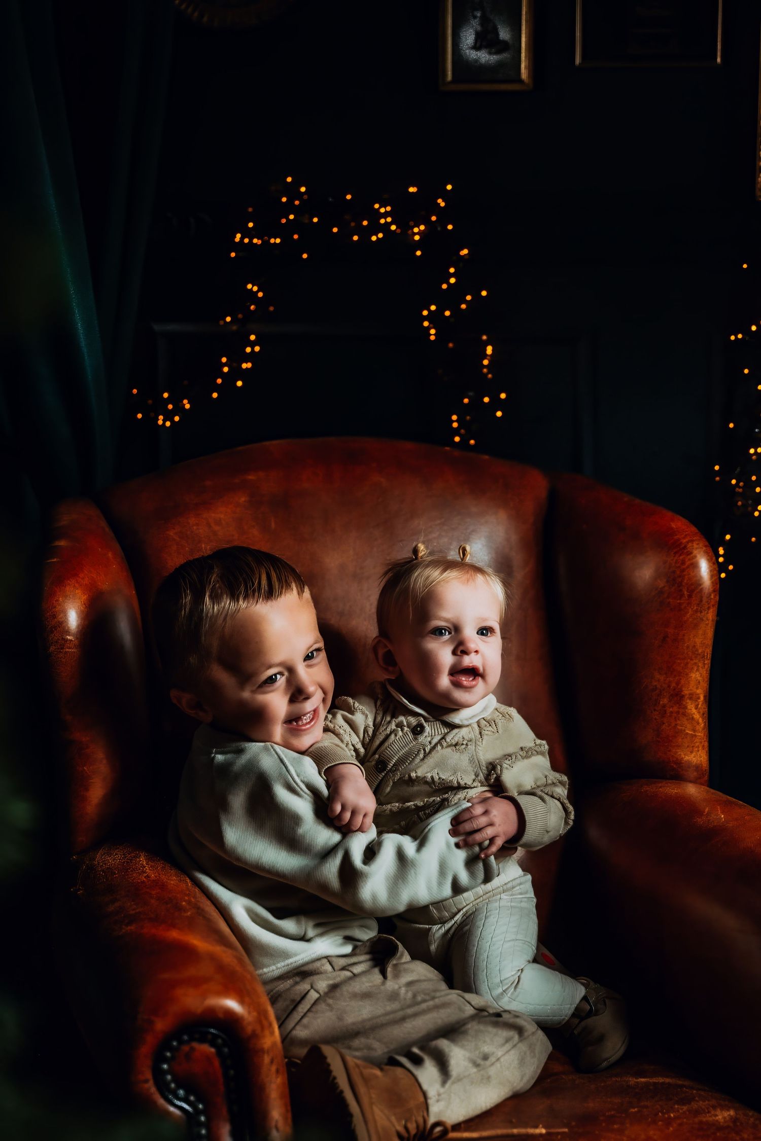 Two small children sit together on a brown leather couch with twinkling lights in the dark background.
