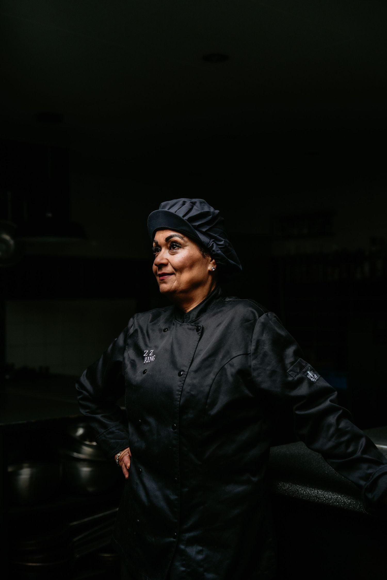 A chef in dark uniform and hat stands in a dimly lit professional kitchen.