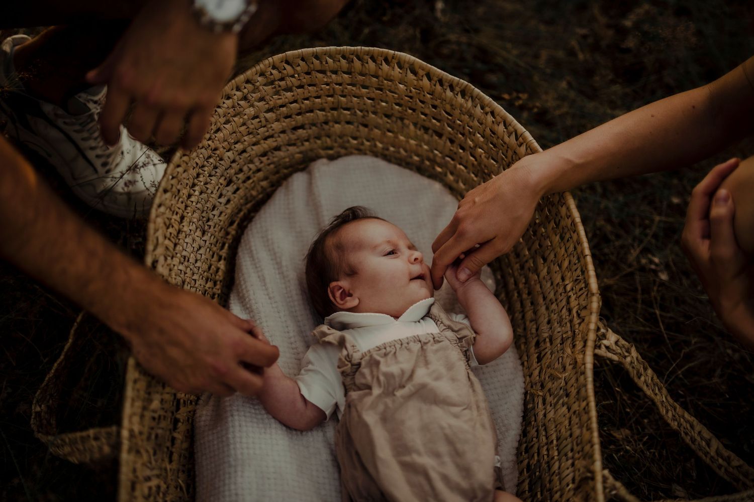 A sleeping baby lies in a woven basket as hands gently reach to touch their thighs.