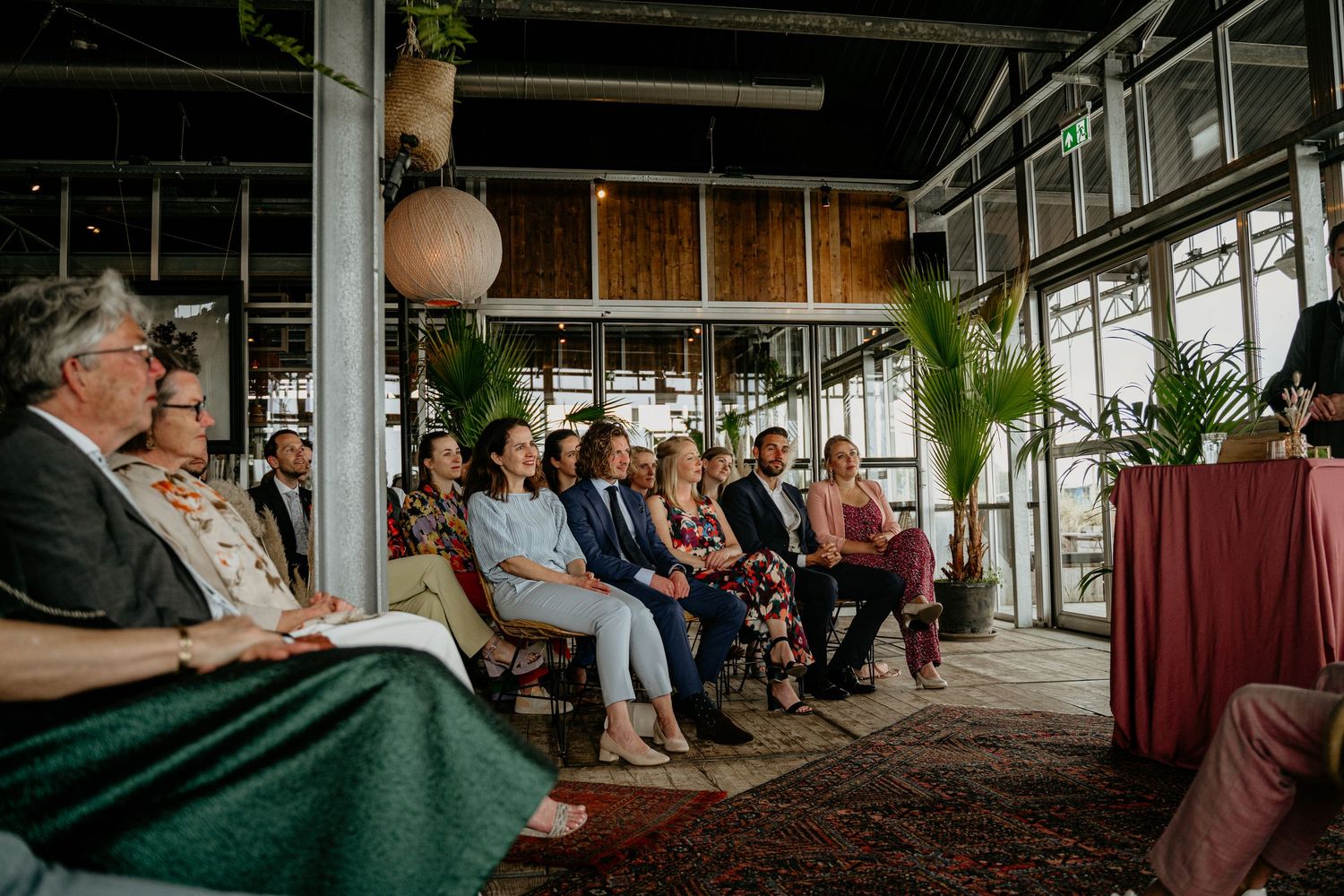 Wedding reception guests seated at tables in an indoor venue with large windows.