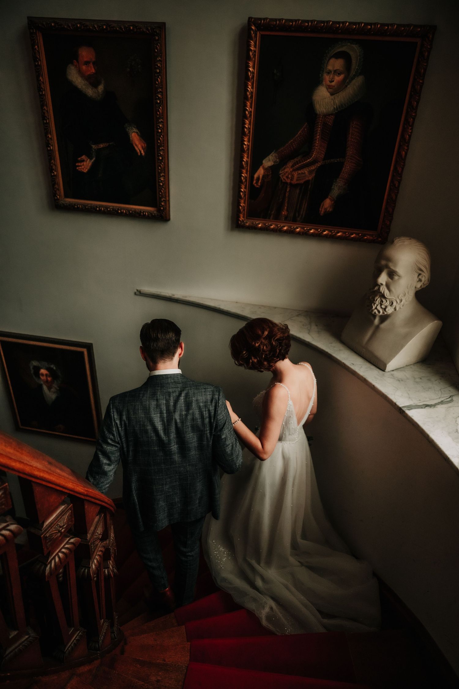 A couple descends an ornate staircase with vintage portraits and a marble bust adorning the walls.