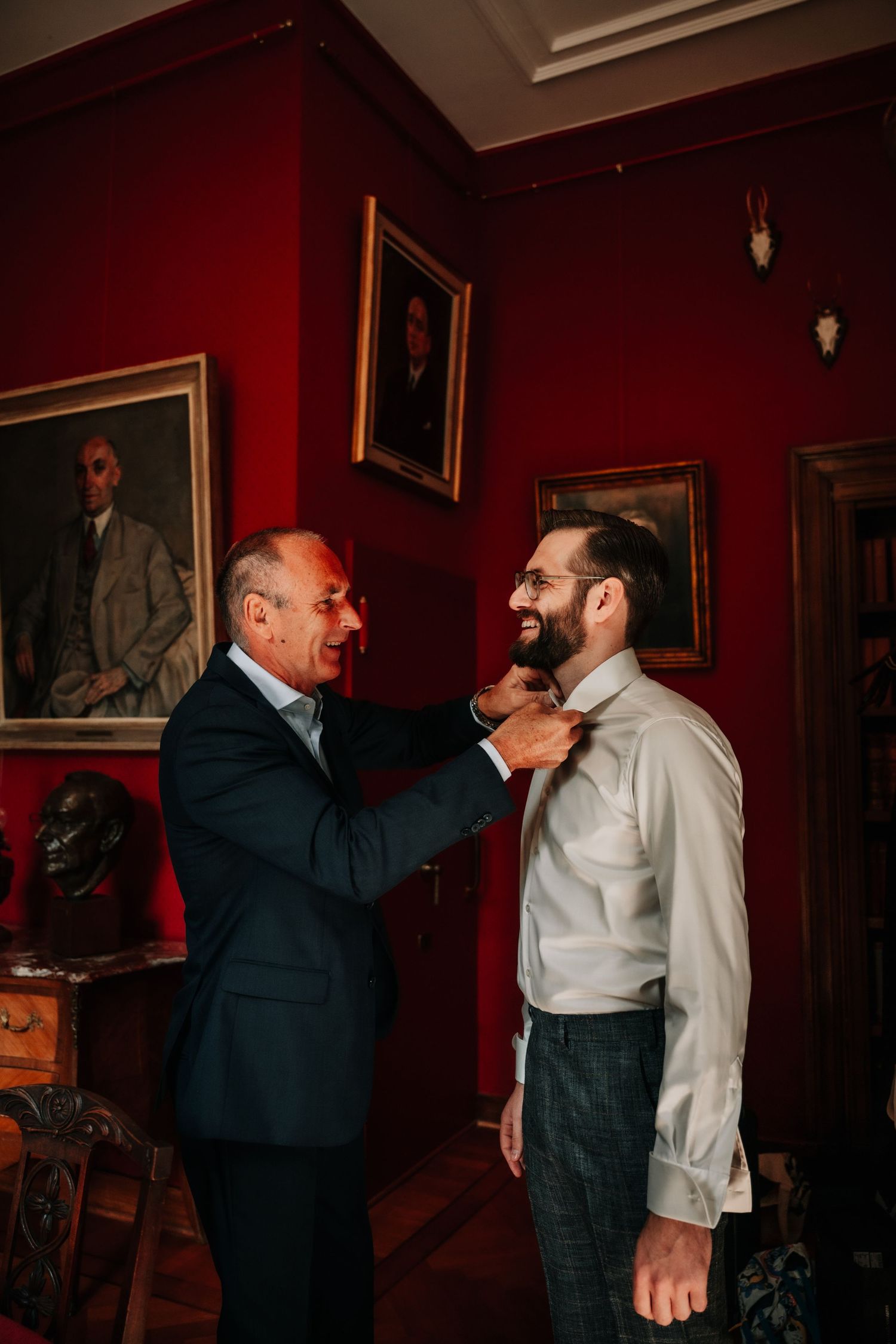 A wedding preparation scene in a room with red walls showing adjustments being made to formal attire.