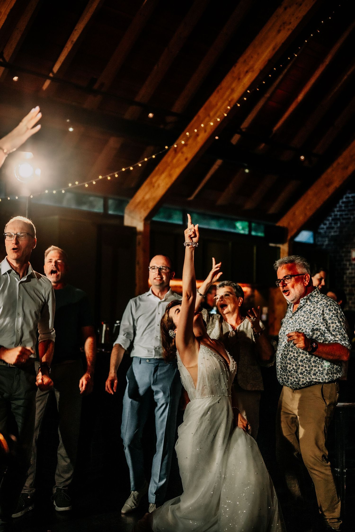 Wedding guests dance together in a rustic barn venue illuminated by warm string lights during an evening reception.