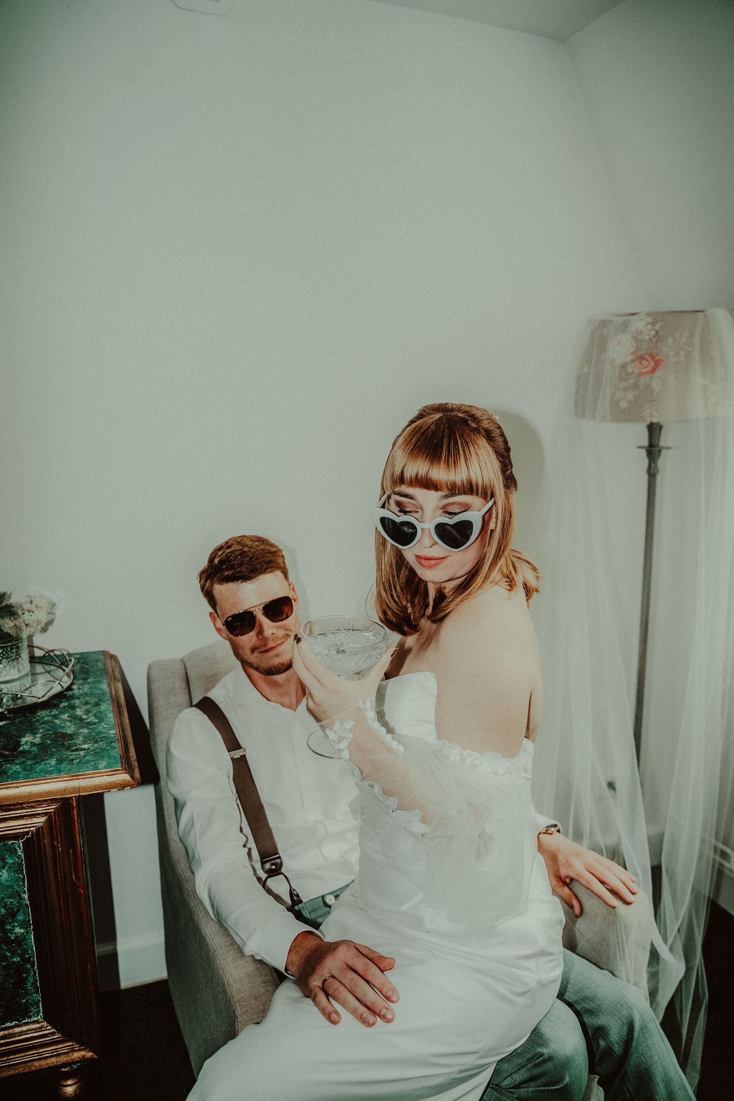A stylish couple wearing sunglasses poses together in a white room with vintage decor.