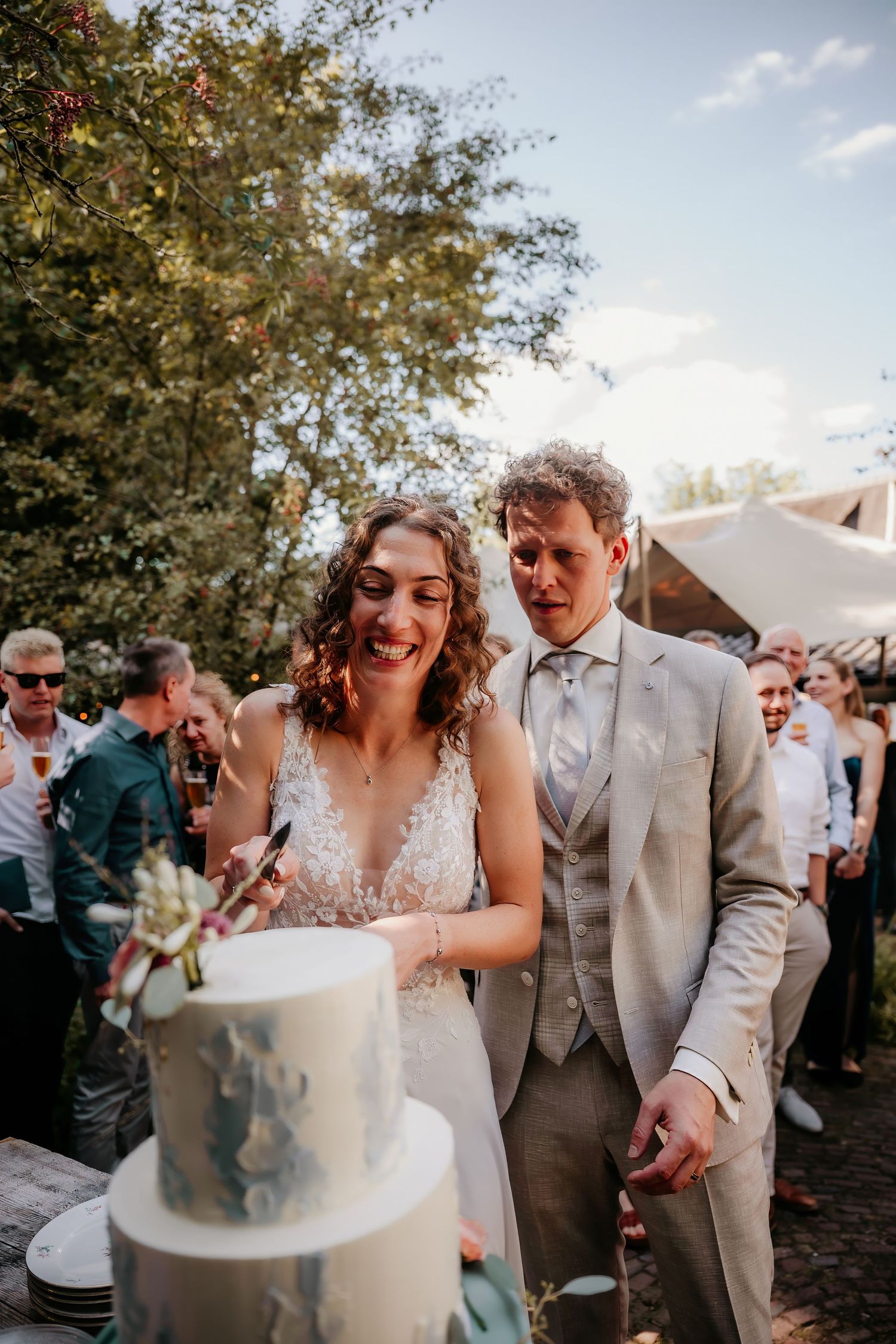 Newlyweds share a joyful moment while cutting their elegant tiered wedding cake at an outdoor reception.